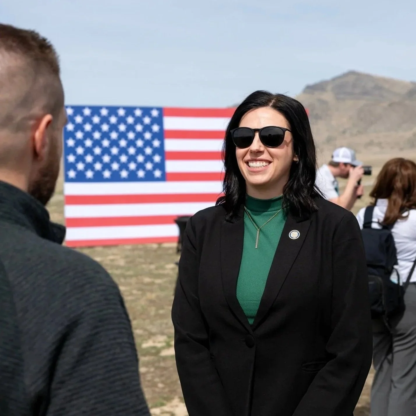 A woman wearing sunglasses and a black blazer smiles at a man with short hair, outdoors with an American flag backdrop and other people in the background, including a person taking a photograph.