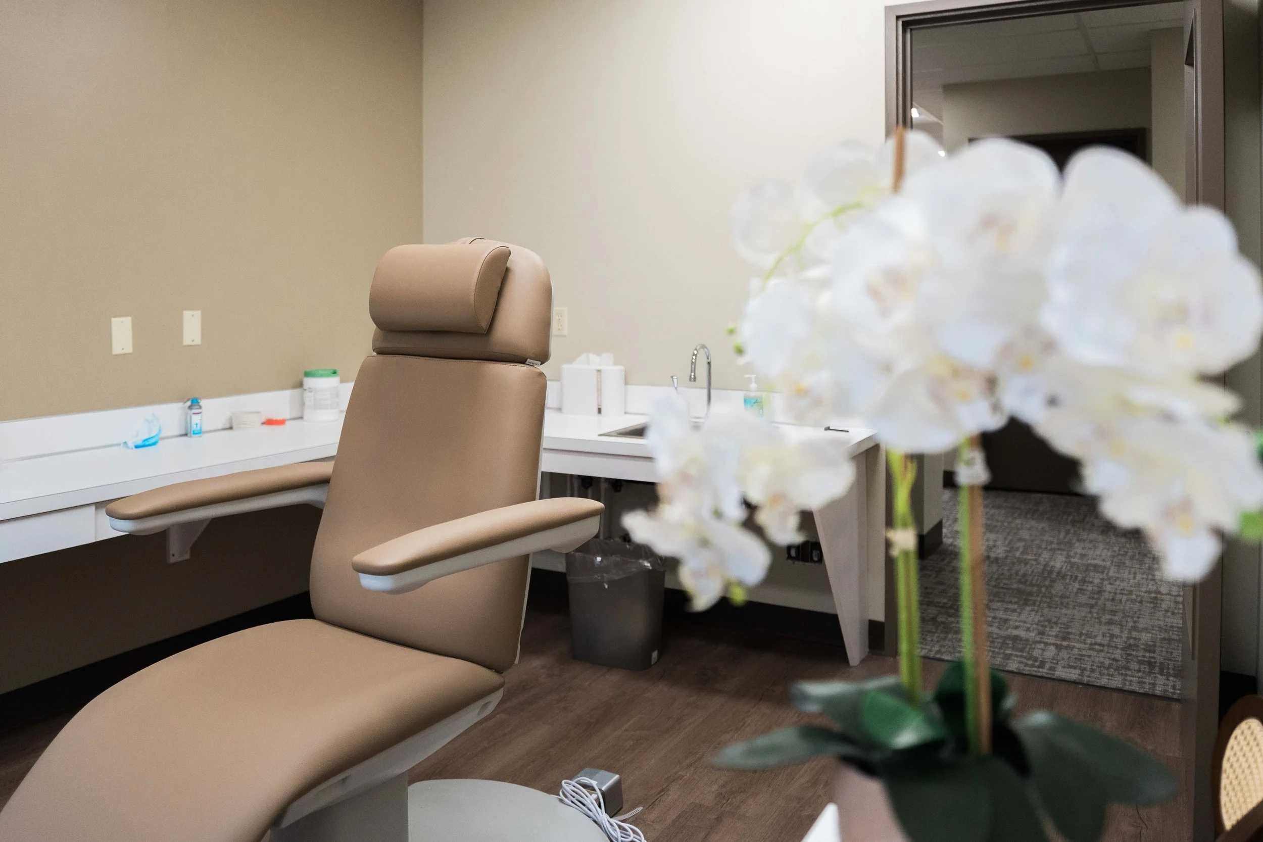 Empty medical or dental exam room with a beige reclining chair, counter with medical supplies, and a sink. A blurry white orchid flower is in the foreground.
