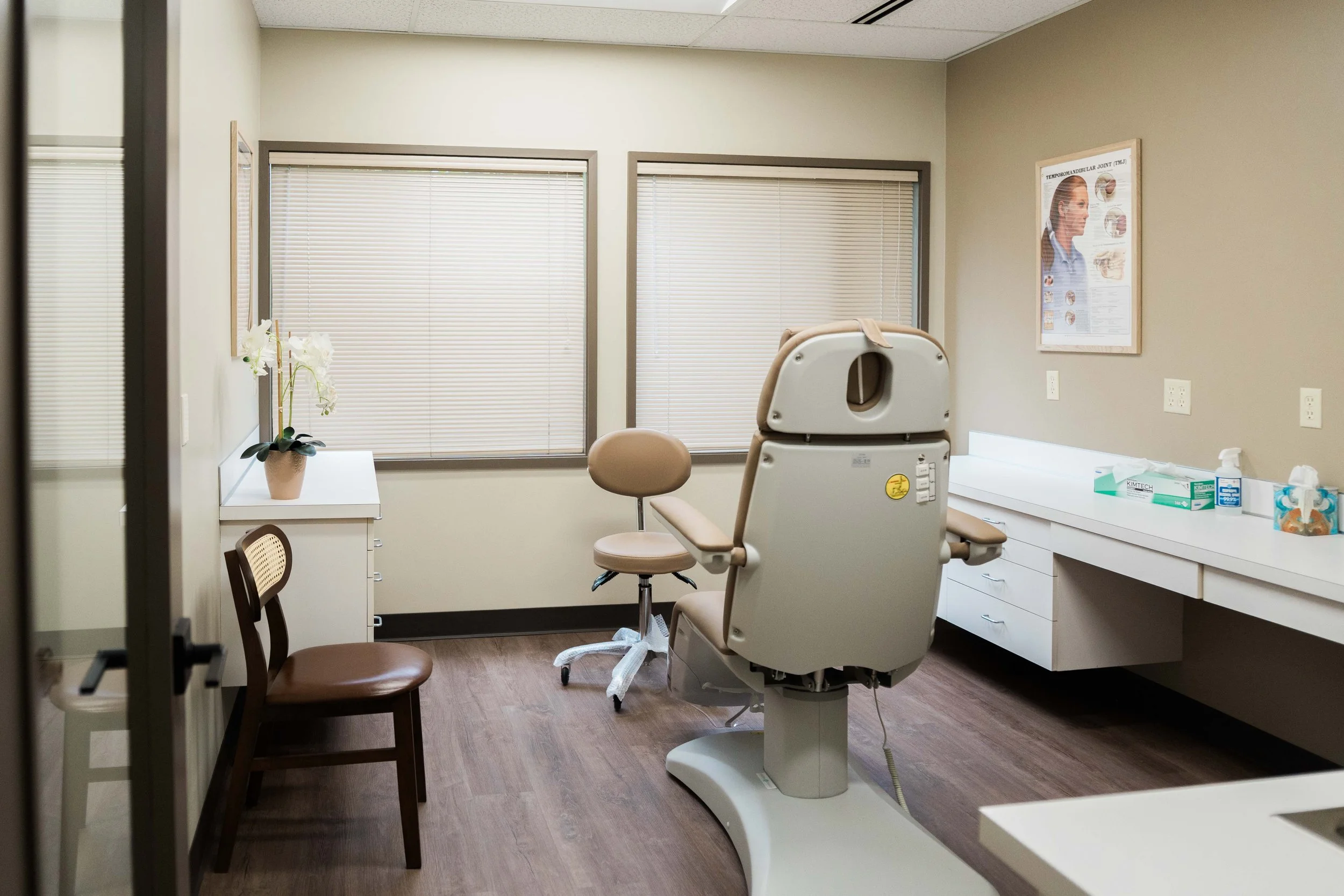 An examination room with a medical chair, two wooden chairs, and a white desk with medical supplies. There is a potted plant on the desk and a medical poster on the wall.
