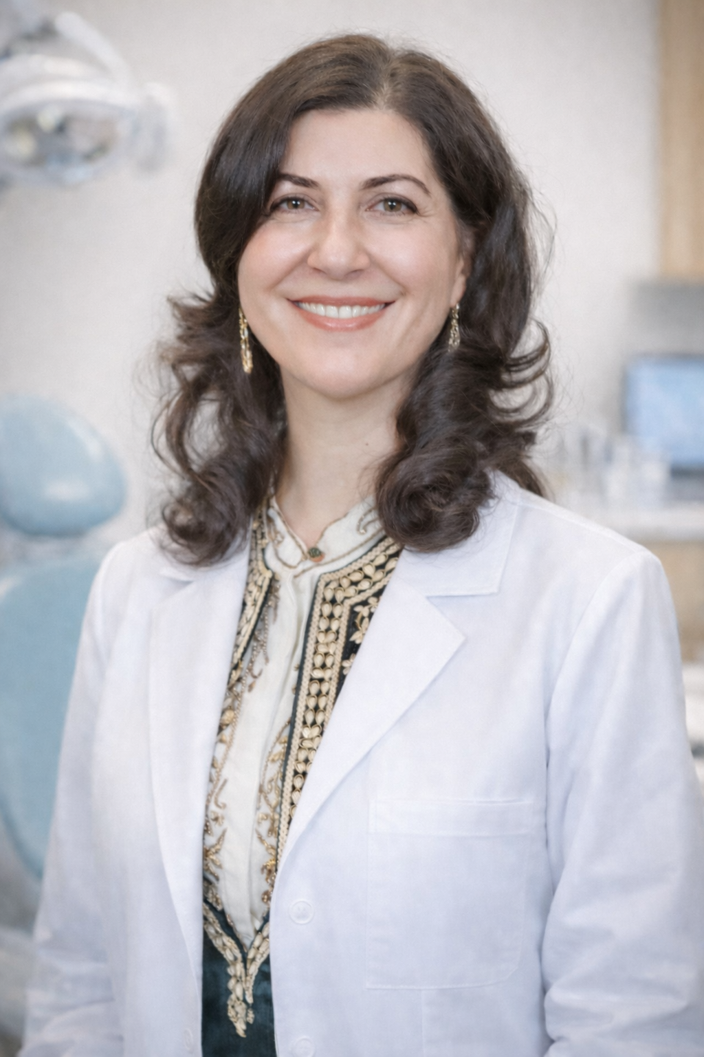 A smiling woman with dark brown, wavy hair, wearing a white lab coat and gold jewelry, standing in a laboratory or medical setting.