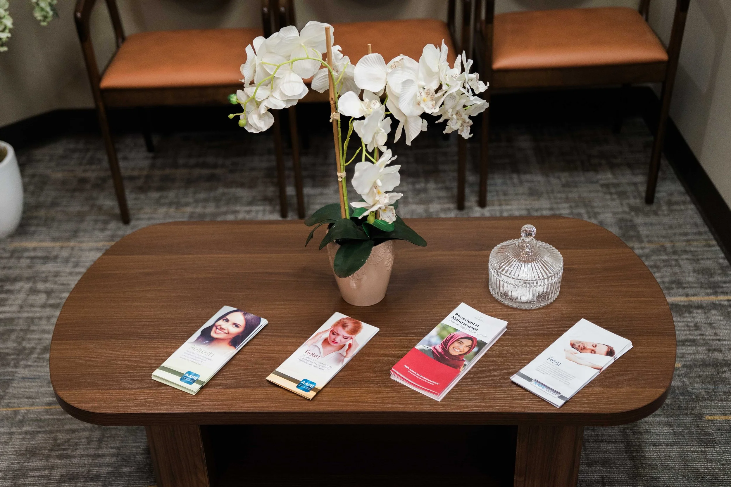 A wooden table with palm-sized brochures about dental health and stress relaxation. A white orchid flower in a beige pot sits at the center of the table, and a decorative glass jar is placed next to it..