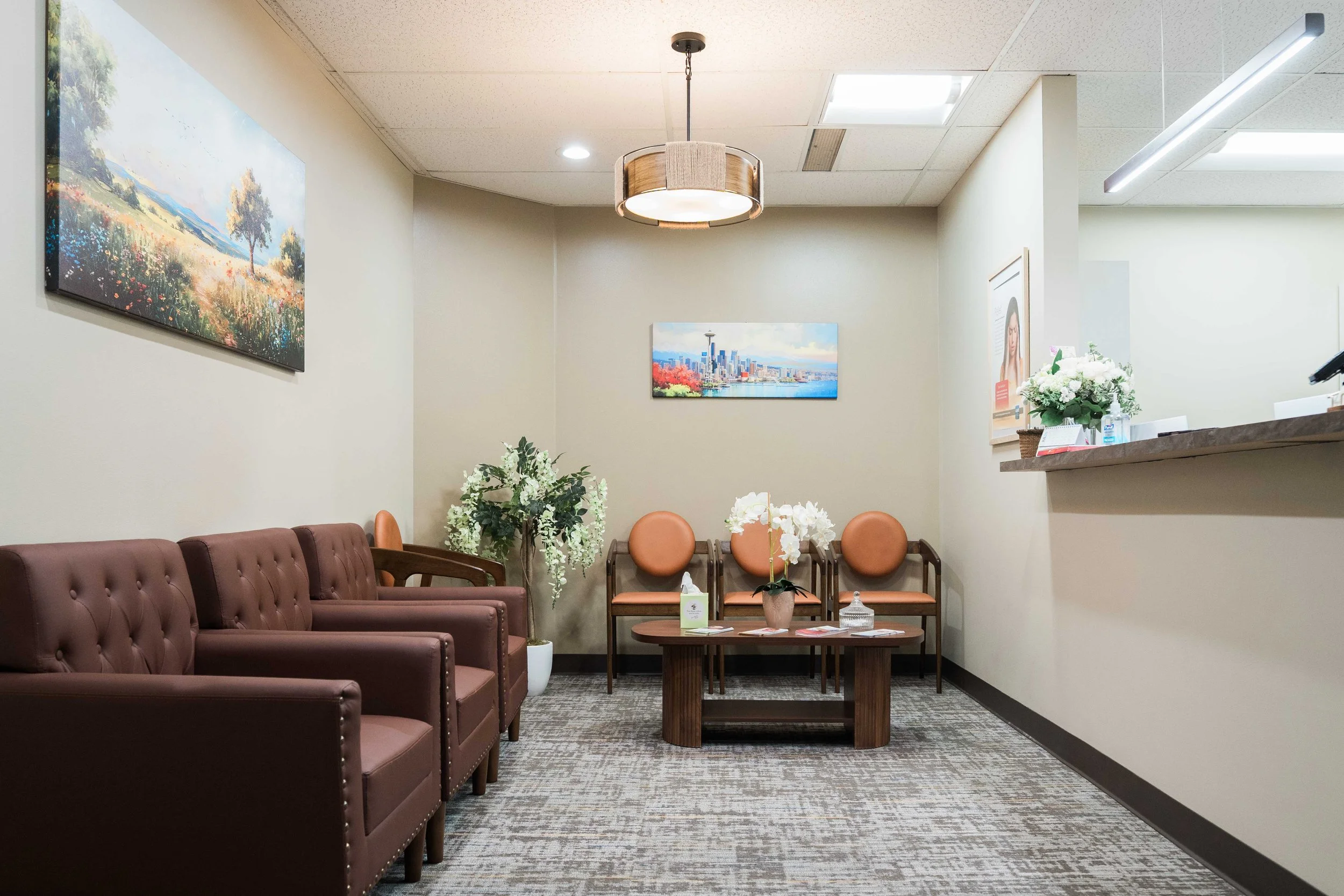 A waiting room with pink couches, orange chairs, a wooden coffee table, a potted plant with white flowers, and artwork on light-colored walls, illuminated by ceiling lights.