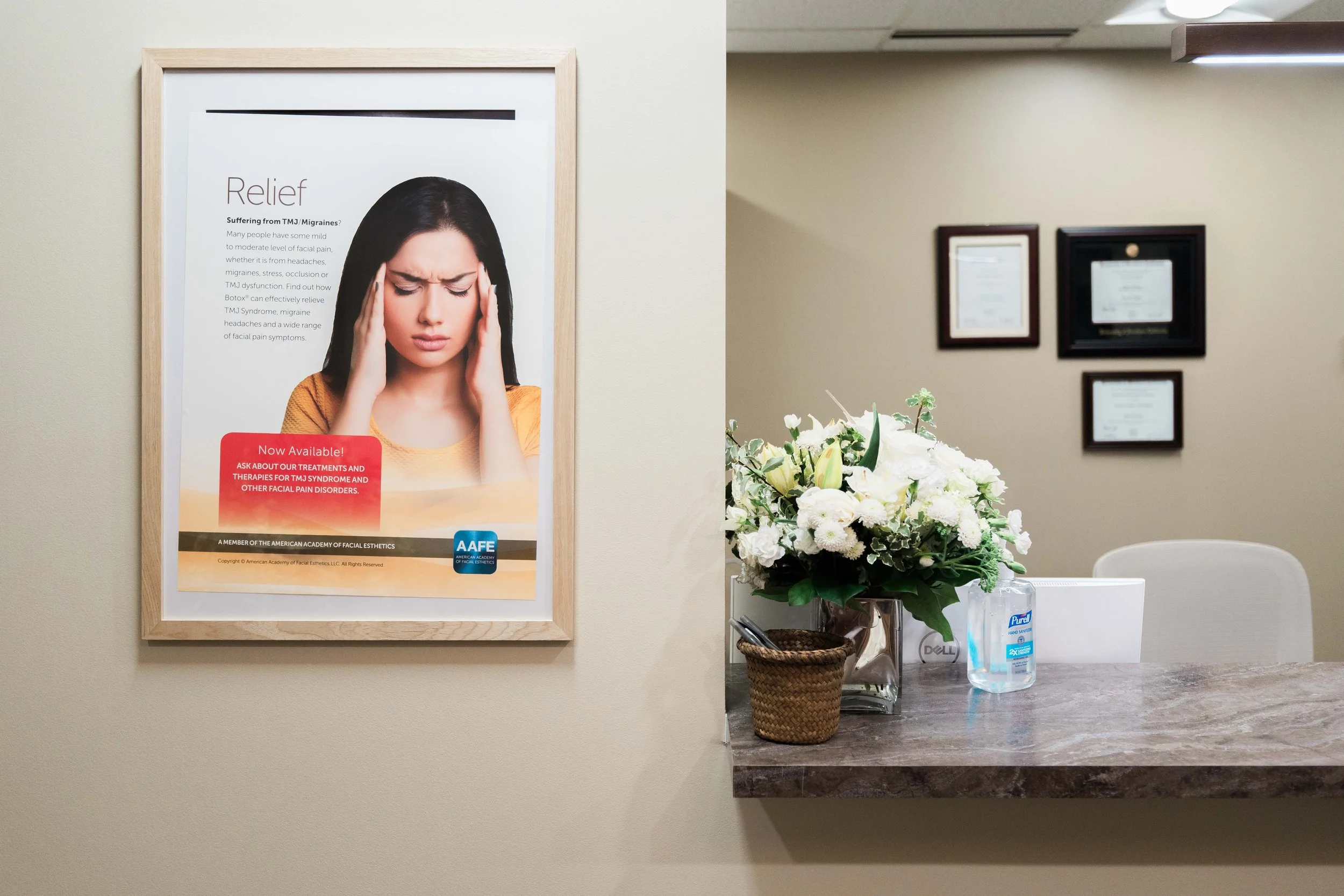 Interior of a medical or aesthetic clinic with a framed health-related poster on the wall, a flower arrangement on the counter, and framed certificates or diplomas on the wall.