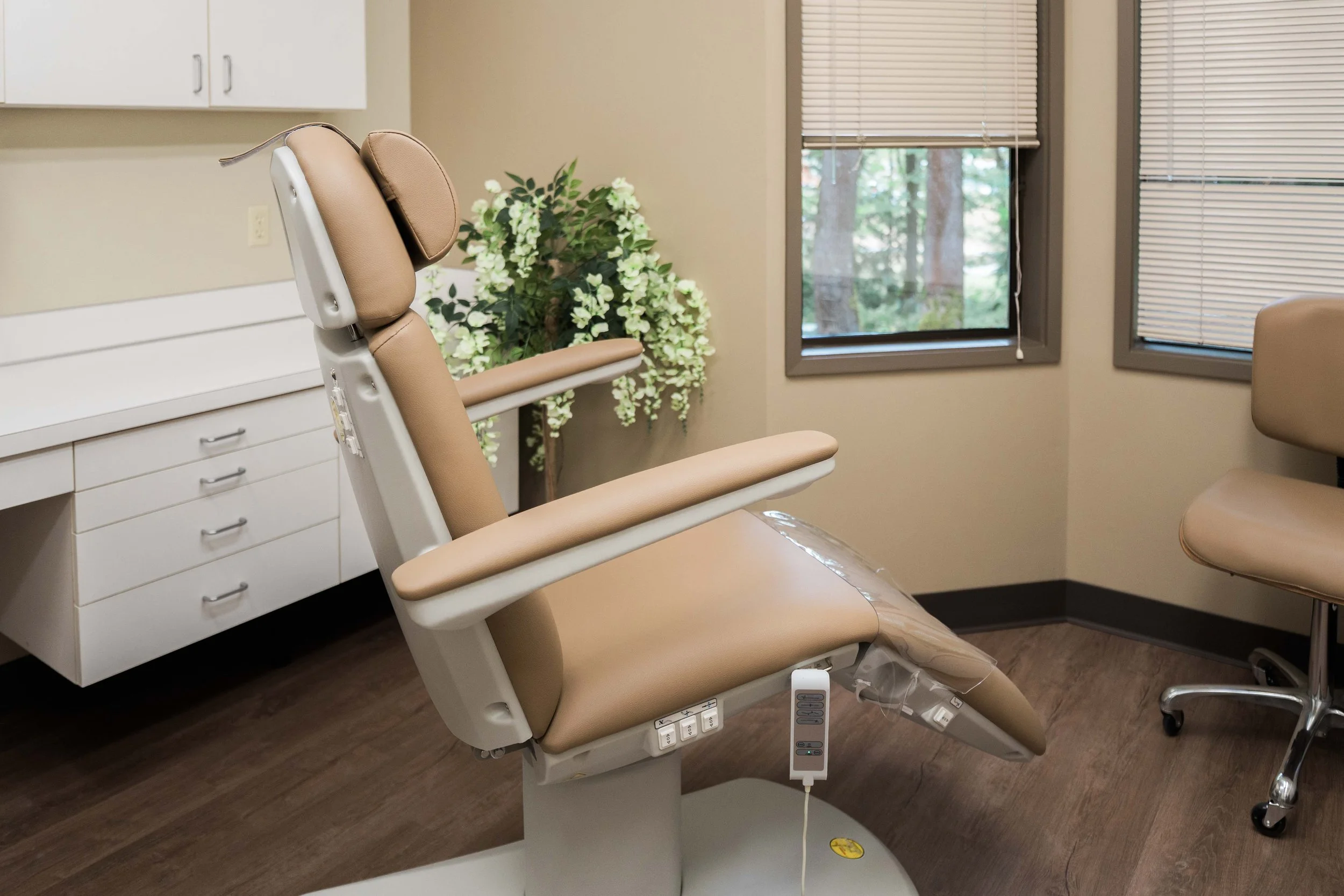 A medical examination room featuring an adjustable beige examination chair positioned in front of a white cabinet and a window with blinds, overlooking trees outside.