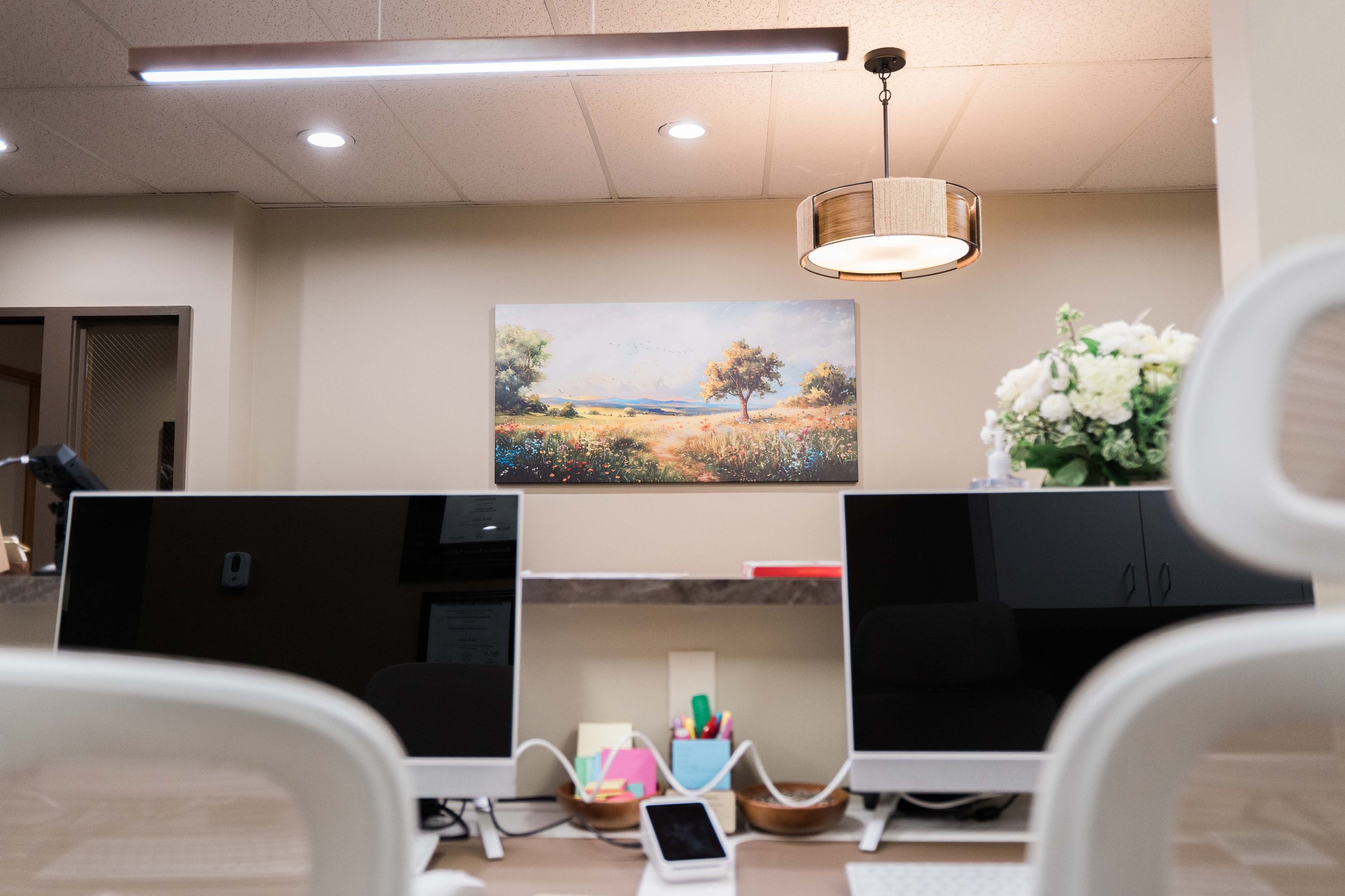 Office reception area with a painting of a landscape on the wall, a modern ceiling light, a bouquet of white flowers, and several computer monitors on desks.
