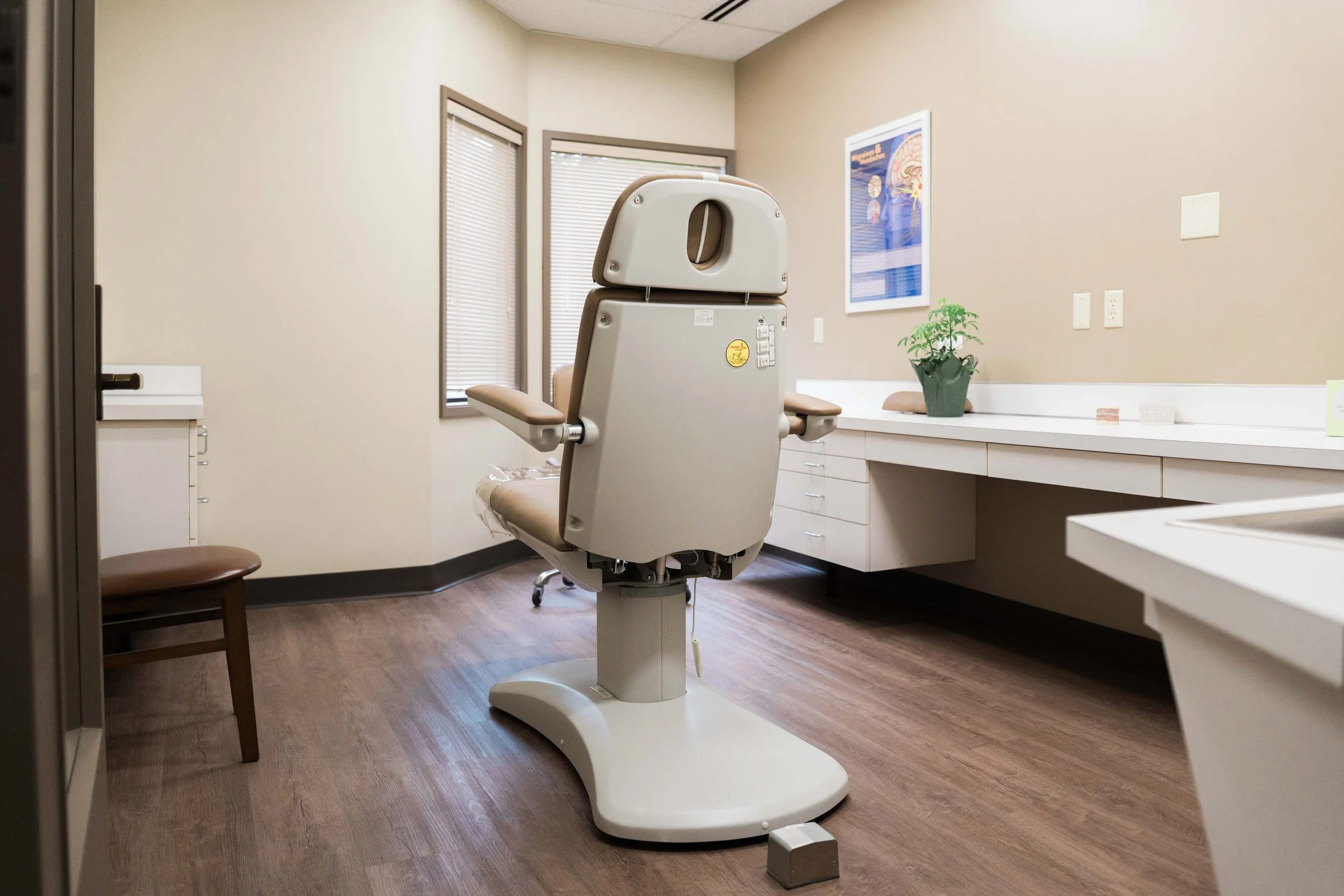 Exam room with medical chair, wooden stool, and medical supplies, window blinds, and wall poster.