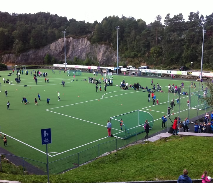 Fotballbane med mange mennesker, noen spiller fotball, andre står og ser på, omgitt av skog og fjell.