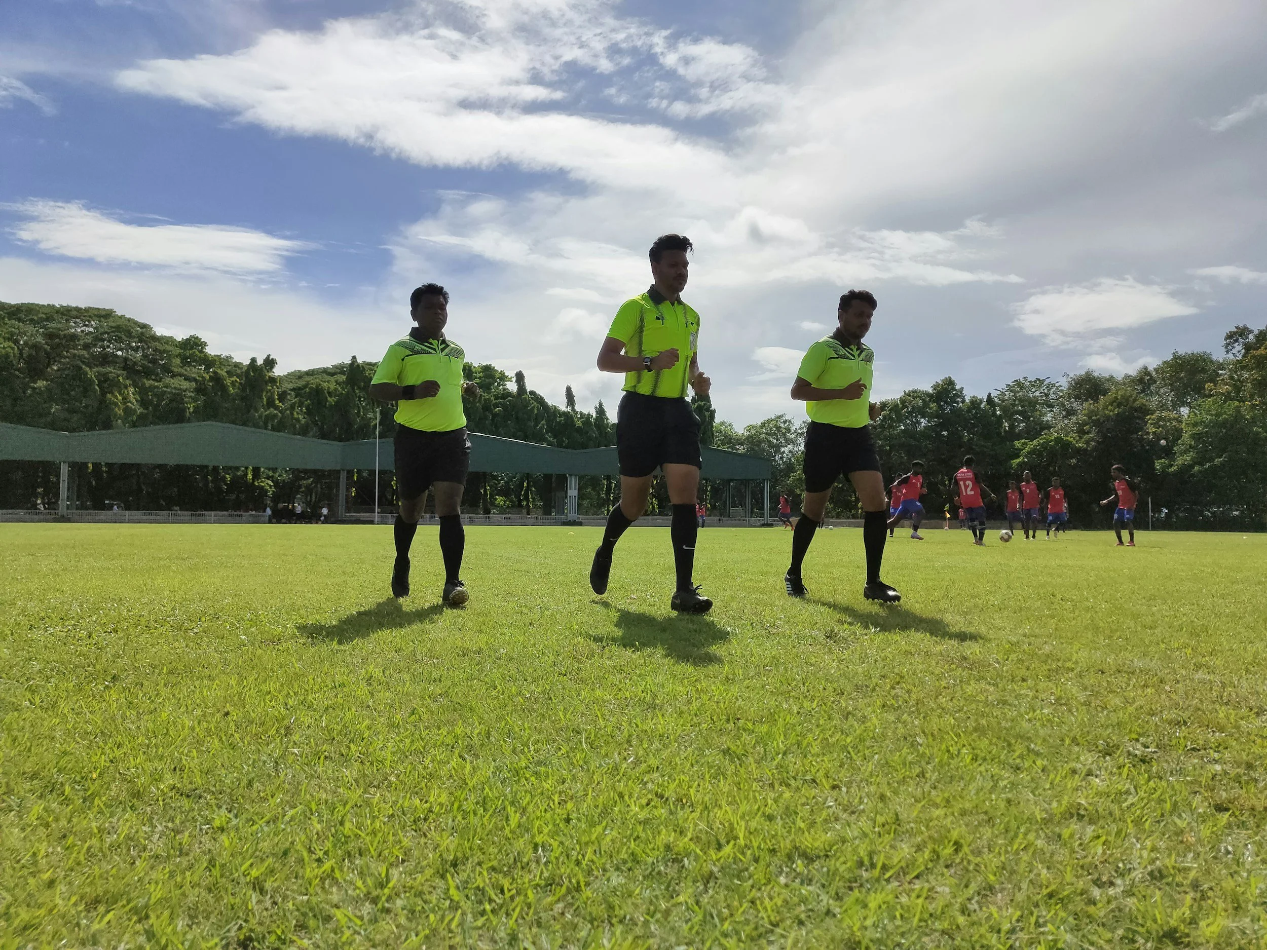Tre fotballspillere løper på en gressbane, iført lysegrønne treningstøy, med flere spillere i røde trøyer i bakgrunnen, under en delvis overskyet himmel.