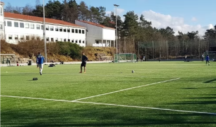 Fotballbane med personer som trener, omgitt av gjerd og bygninger i bakgrunnen på en solrik dag.