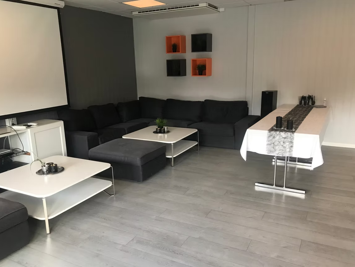 Stue med svart sofa, to hvite bord, veggdekor med fire hyller, og et avlangt bord med hvit duk og svart dekor.