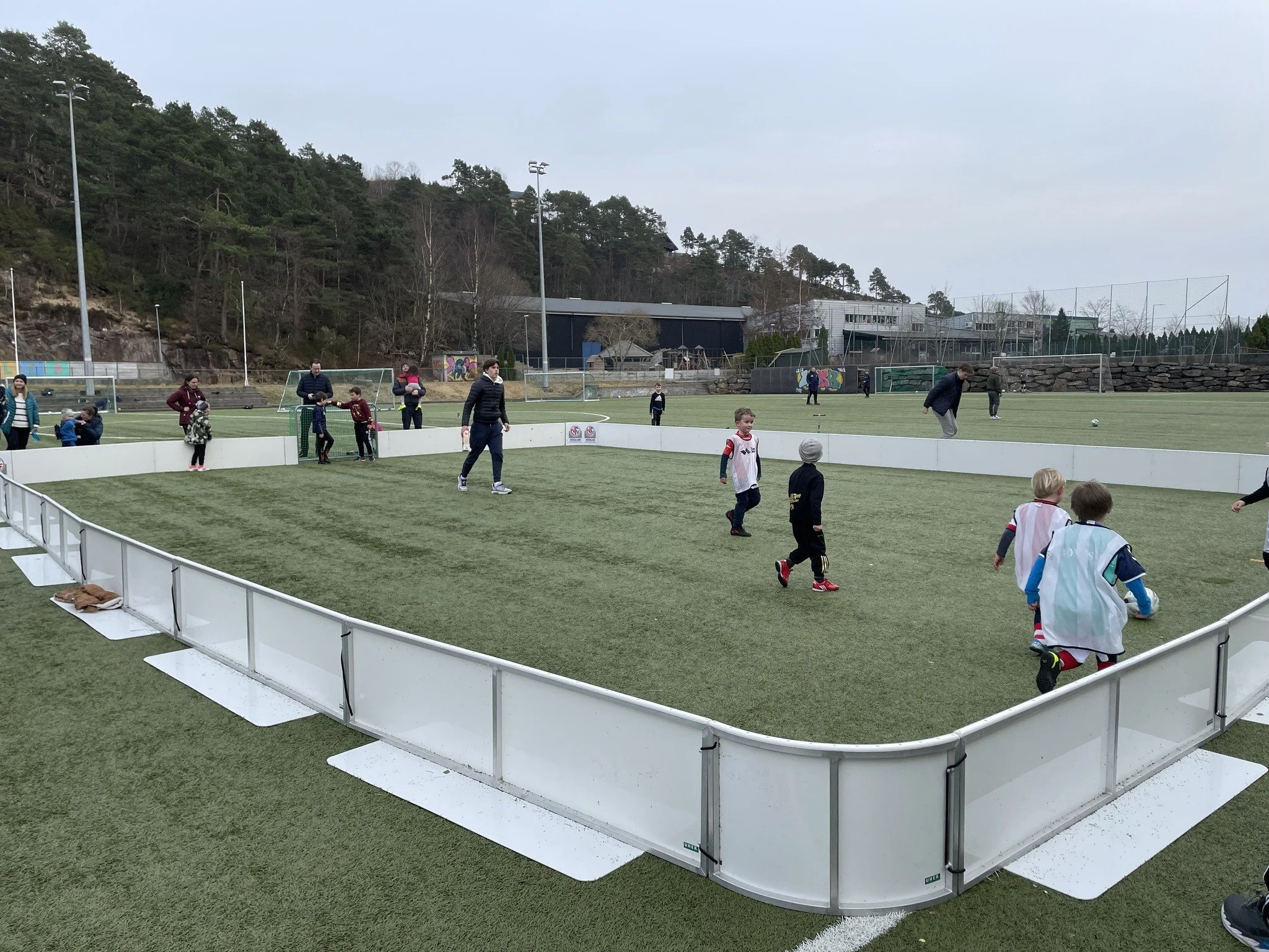 Barn og voksne på en kunstgressbane, flere barn spiller fotball, omgitt av midlertidig barriere og med trær og bygninger i bakgrunnen.