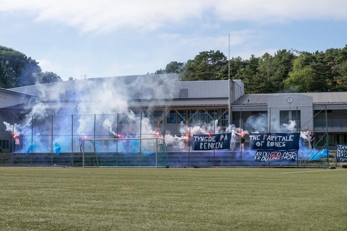 Fotballbane med brannfarlig røyk og blå folkeflagg, som viser protester med bannere med teksten "Tyngde på benken" og "The fairytale of bones".