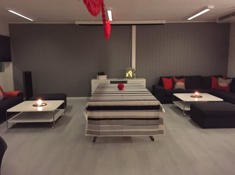 Et rom med en hvit borddekk med striper og en rødfarget tennisball, omgitt av to sort sofaer med puter, to lave bord med lys, og en svart høyttaler.