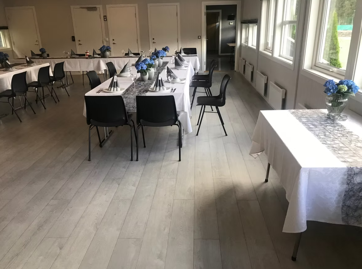 Møterom eller spisesal med bord dekket for en anledning, med hvite duker, blomsterdekorasjoner med blå og hvite blomster, og plass til gjester.