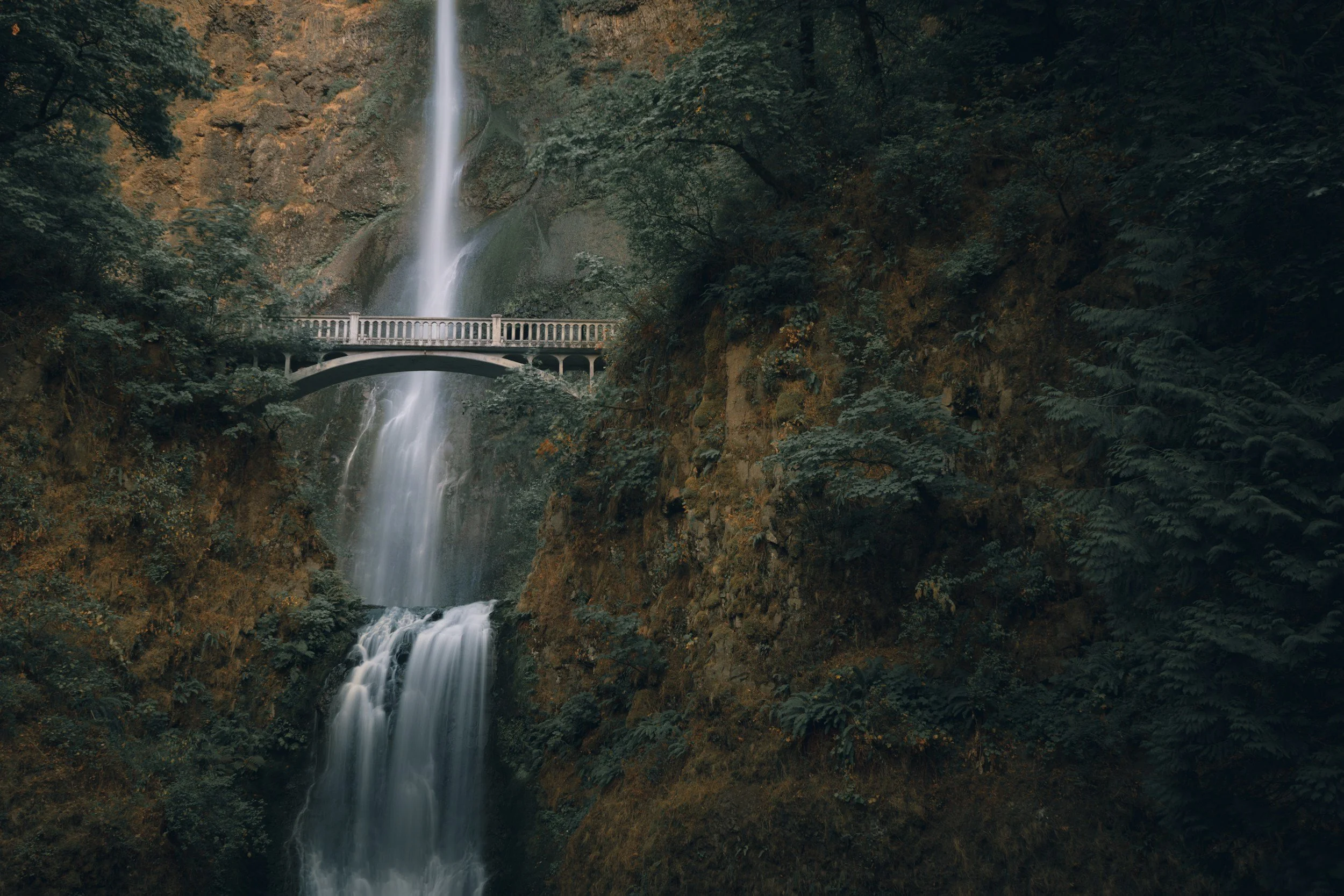 A multi-tiered waterfall flowing down a rocky cliff surrounded by dense green foliage, with a small bridge crossing the waterfall at the middle level.
