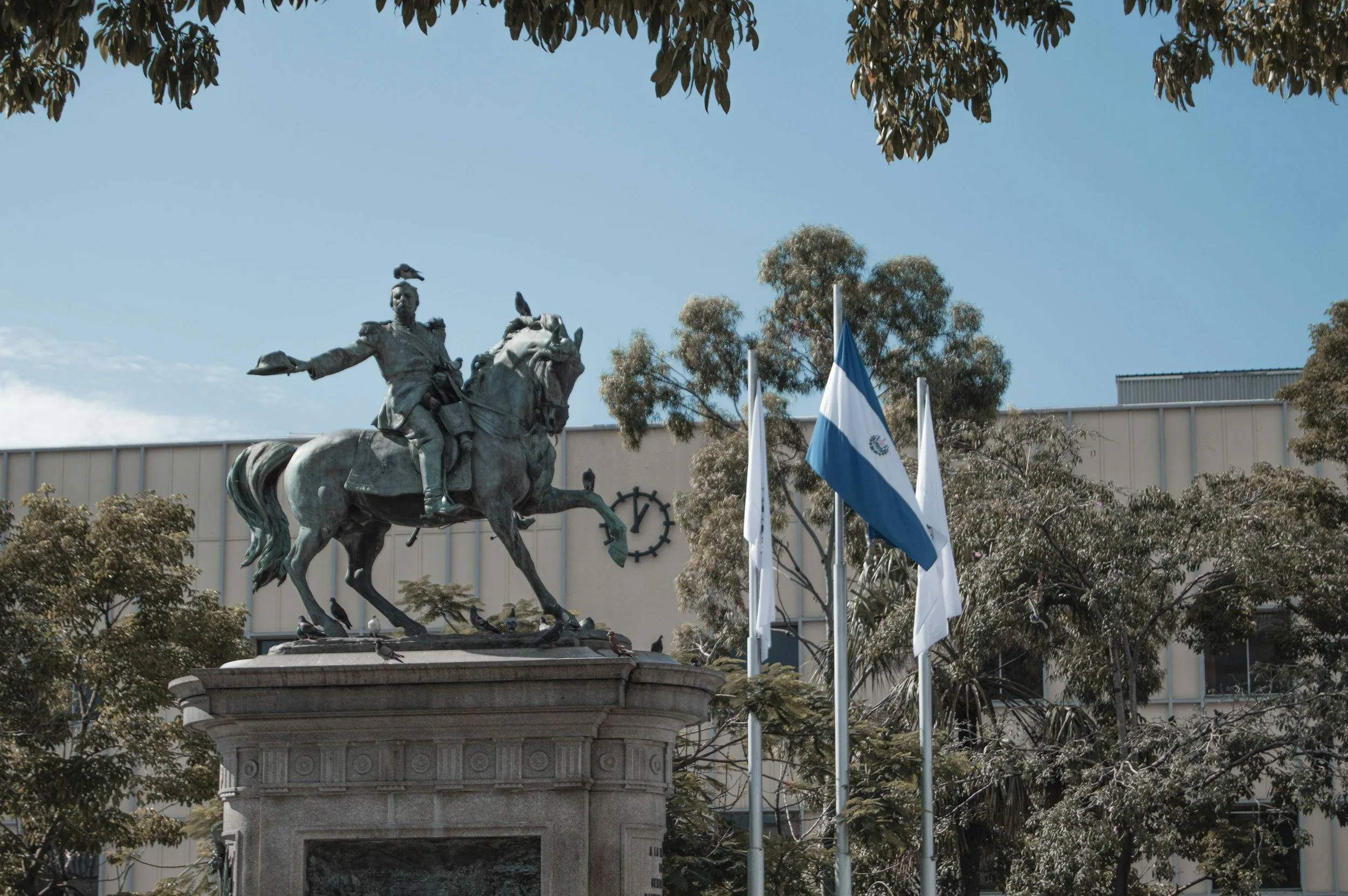 Statue of a man on horseback with outstretched arm, surrounded by flags, trees, and a building in the background.