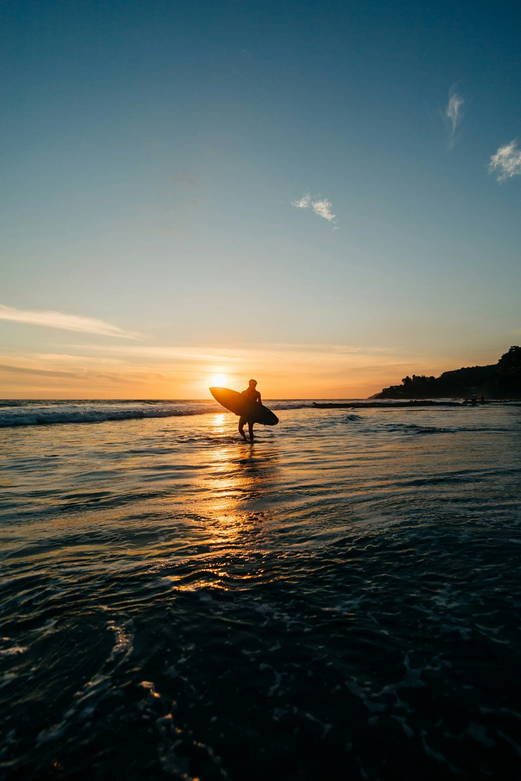 A person carrying a surfboard walking in the ocean during sunset, with a hilly coastline in the distance.