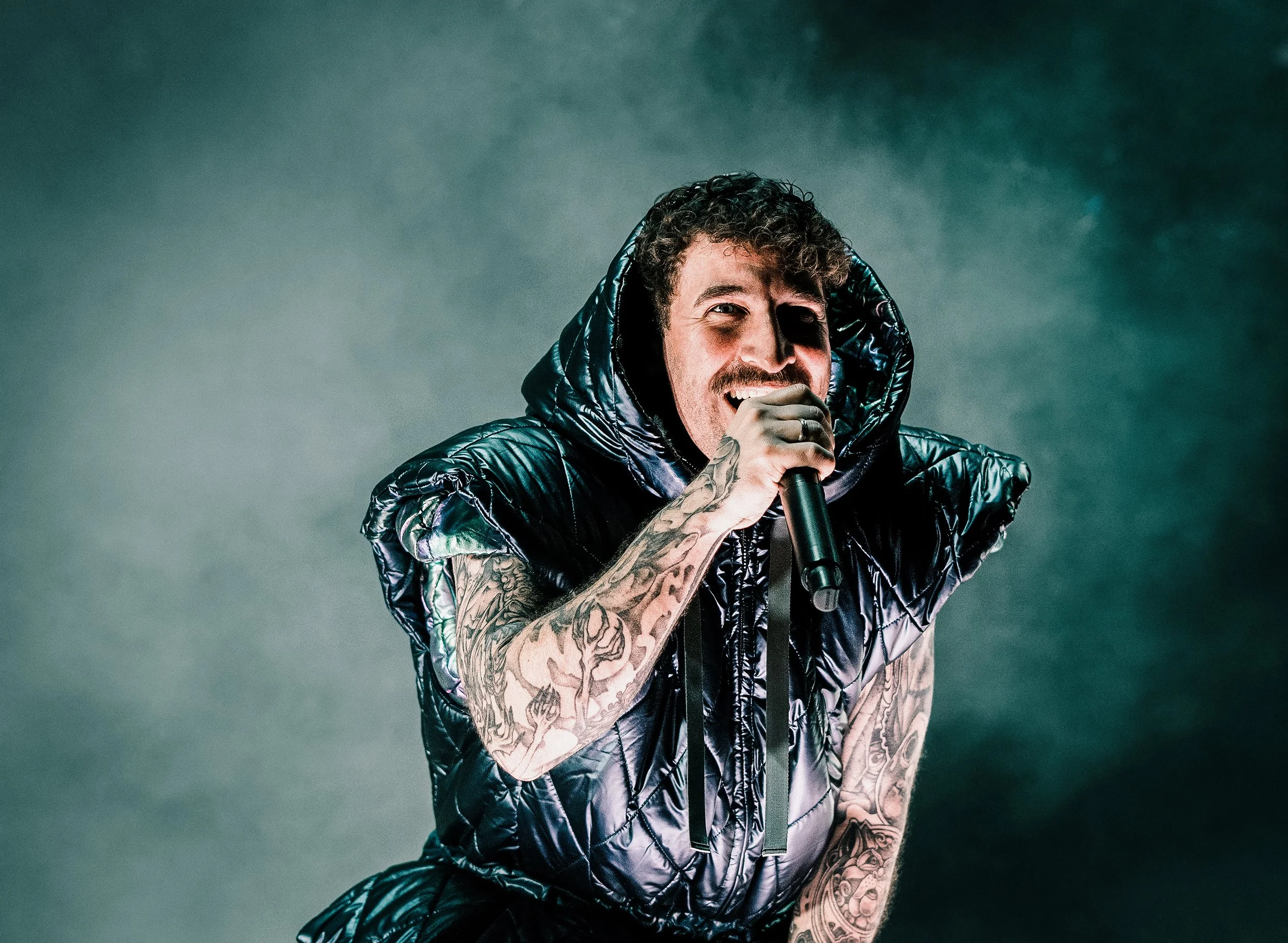A man with curly hair and tattoos on his arms, wearing a shiny black puffer jacket and gray gloves, singing into a microphone against a dark, smoky background.