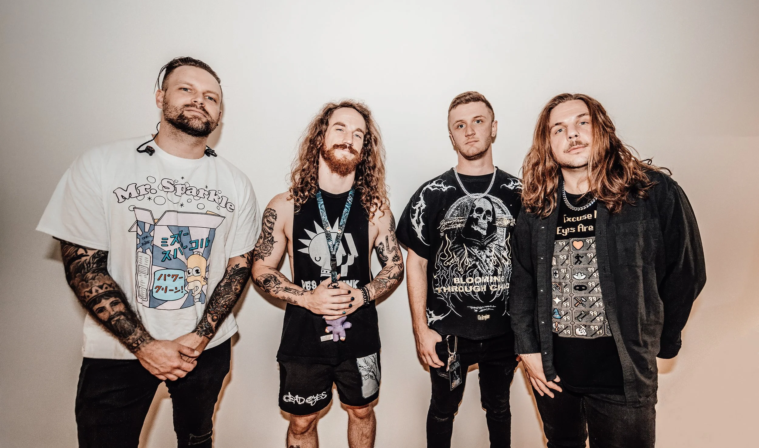 Four men standing against a plain white wall, dressed in casual and band-style clothing, with tattoos and long hair, posing for a photo.
