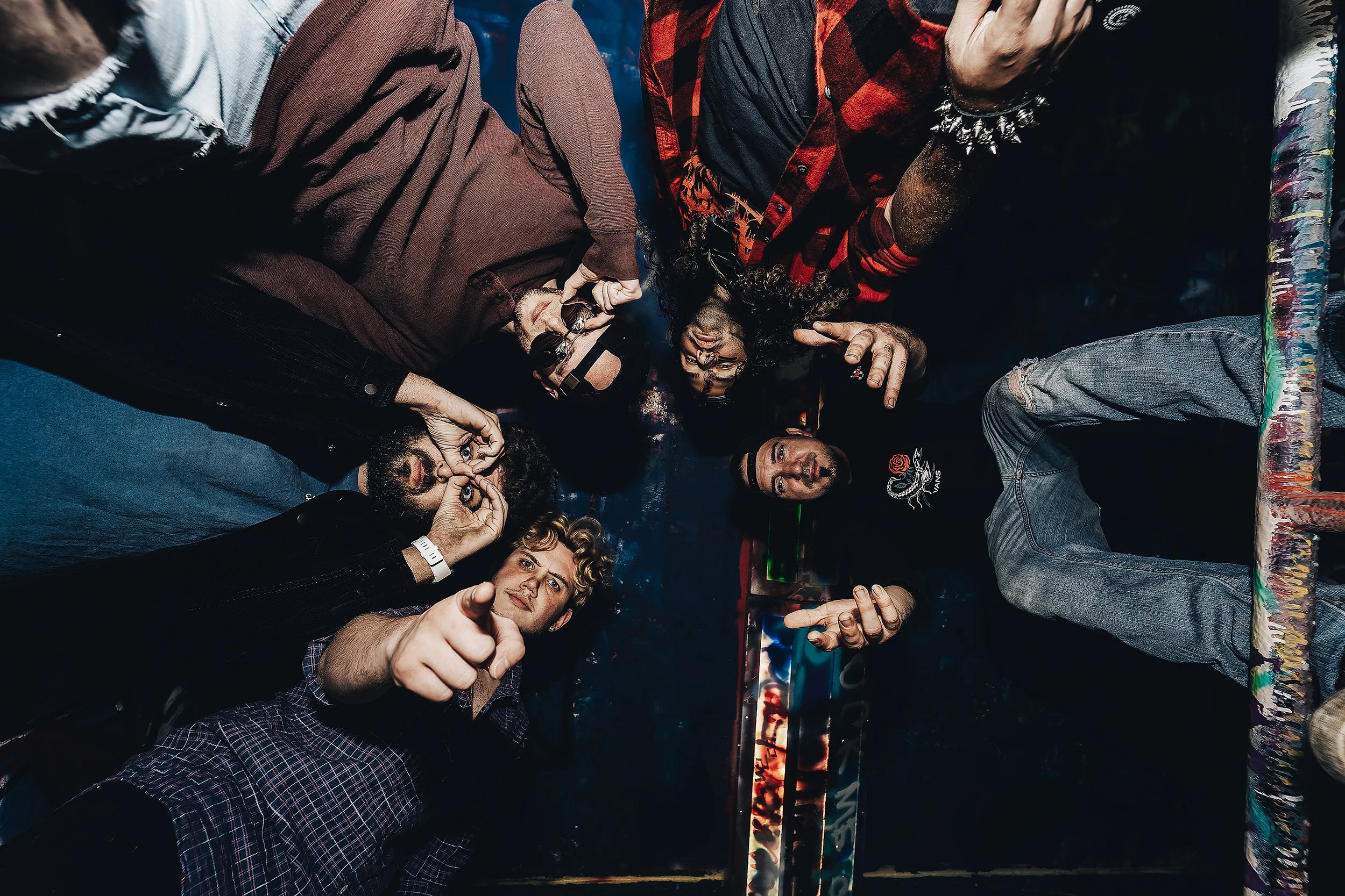 Group of people at a concert or event, some making peace signs, one person pointing directly at the camera, in a dimly lit environment with colorful lighting.