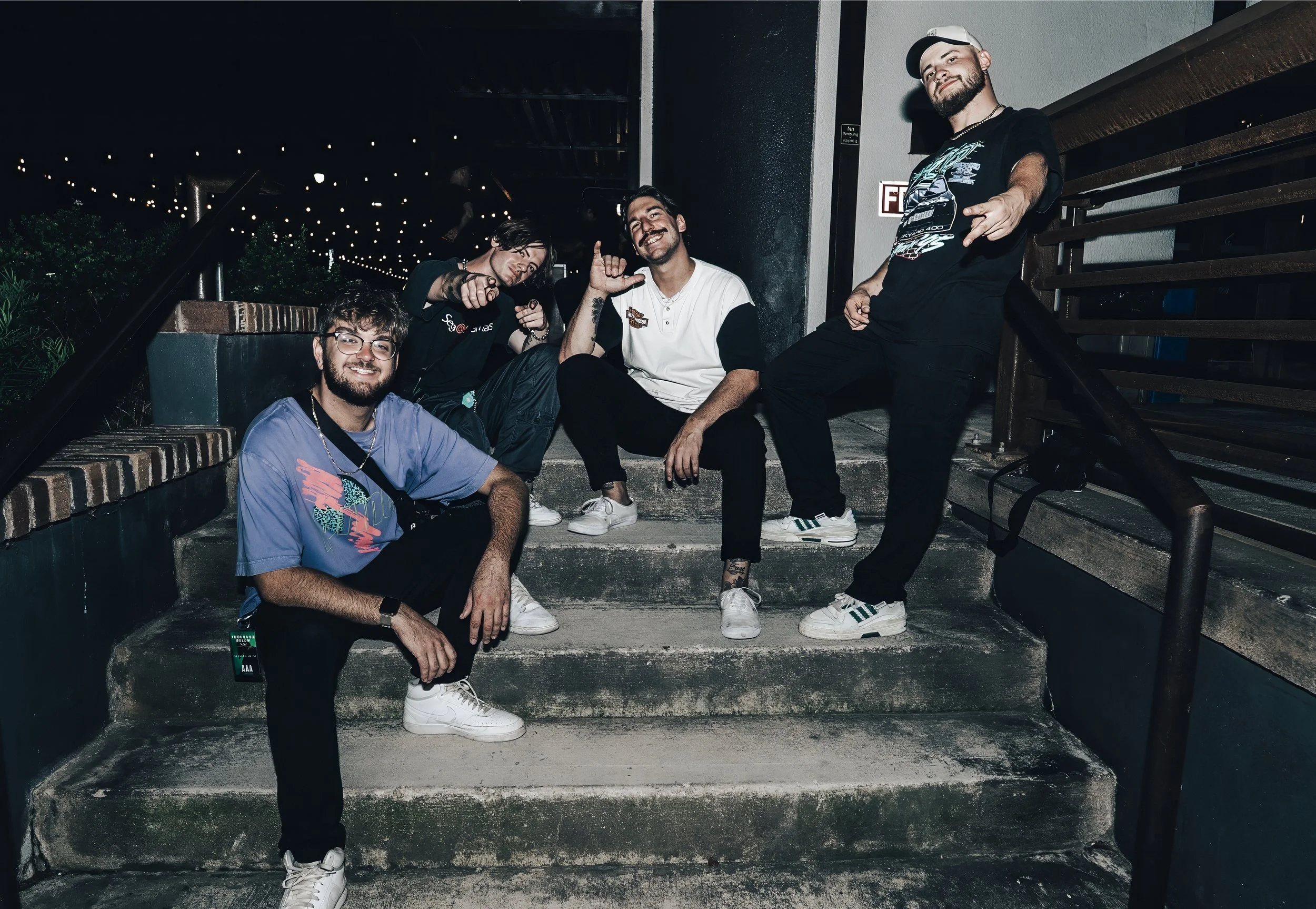 Group of five young men sitting and standing on outdoor concrete stairs at night, smiling and making gestures, with string lights overhead and a wall with a 'Fire Exit' sign behind them.