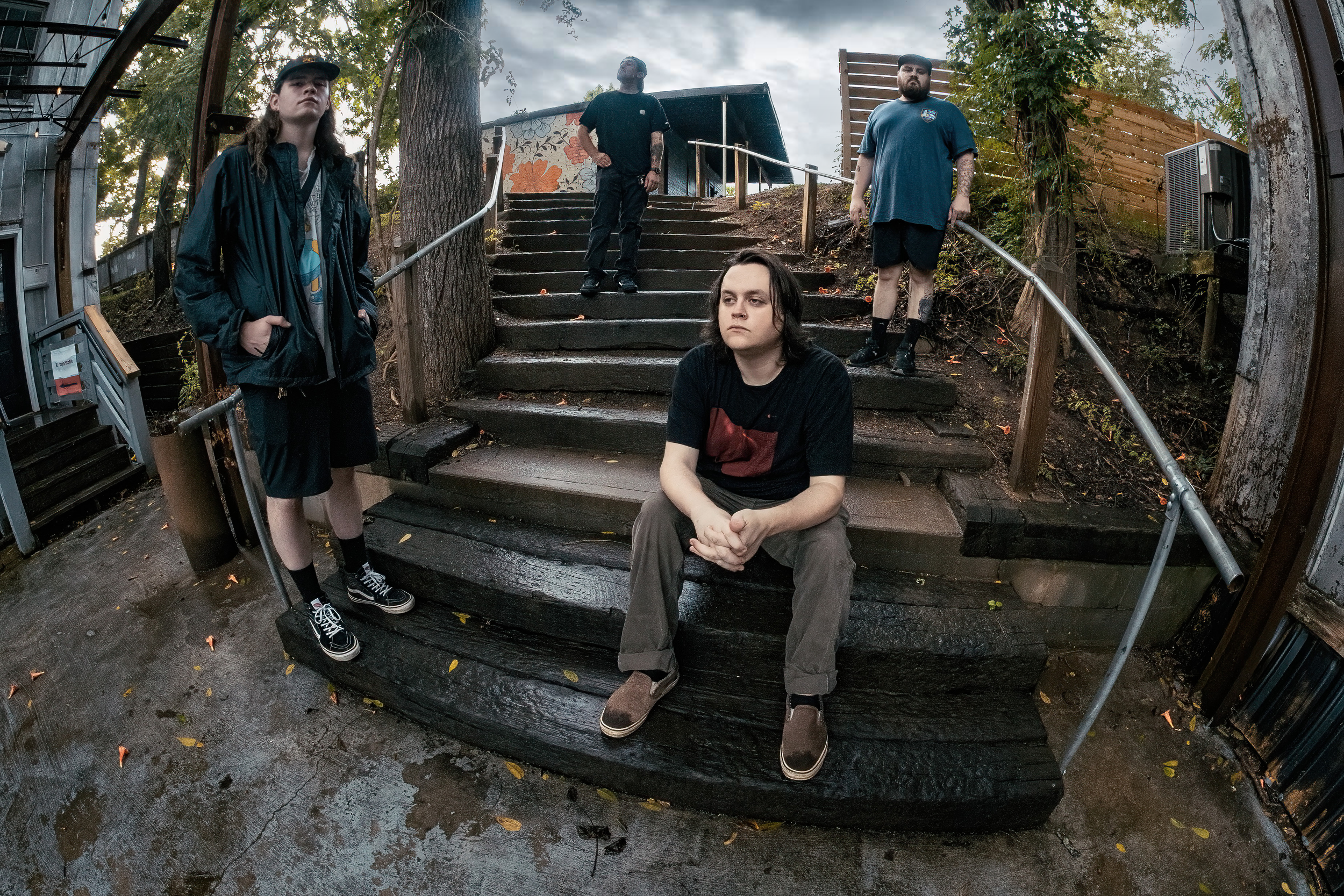 Five young men with tattoos in casual clothing pose on a set of outdoor stairs during cloudy weather. One is sitting on the bottom step, three are standing on the stairs, and one is standing at the top. The surroundings include trees, a wooden fence,
