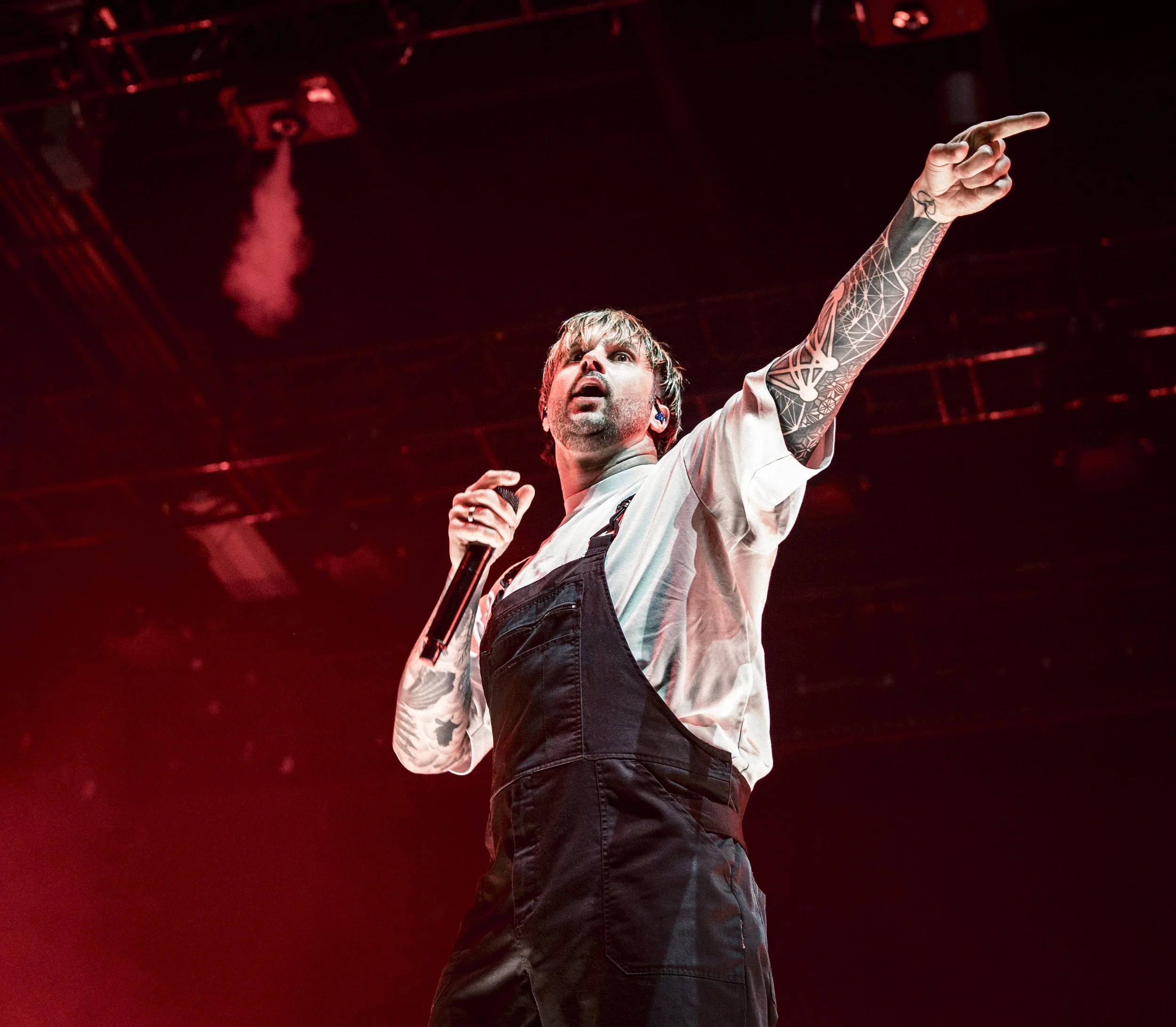 A male performer with tattoos on his left arm, holding a microphone, pointing upward with his right arm, wearing a white shirt and black pants, performing on stage with red lighting.