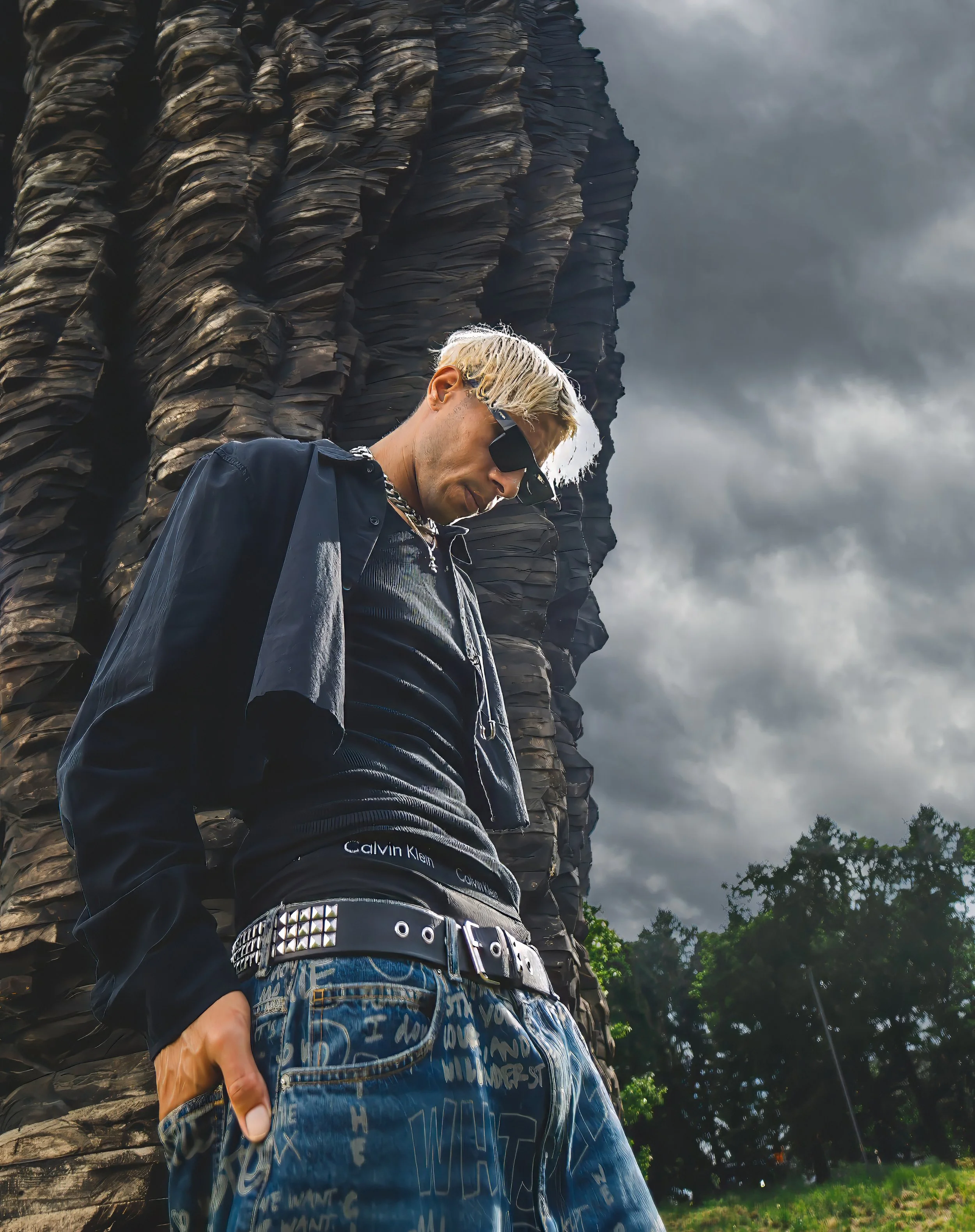 A person with bleached blonde hair and wearing sunglasses, a black jacket, and patterned blue jeans, standing outdoors near a large dark rock formation against a cloudy sky.