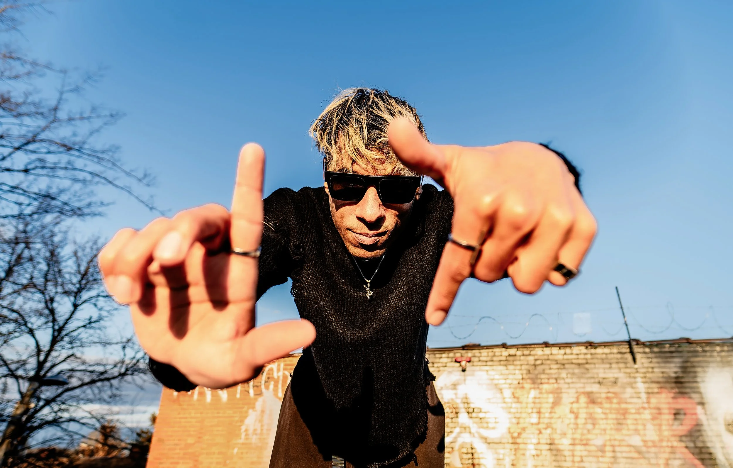 A person wearing black sunglasses and a black top is leaning forward toward the camera, making an framing gesture with their hands. The background shows a clear blue sky, a brick building, and leafless trees.
