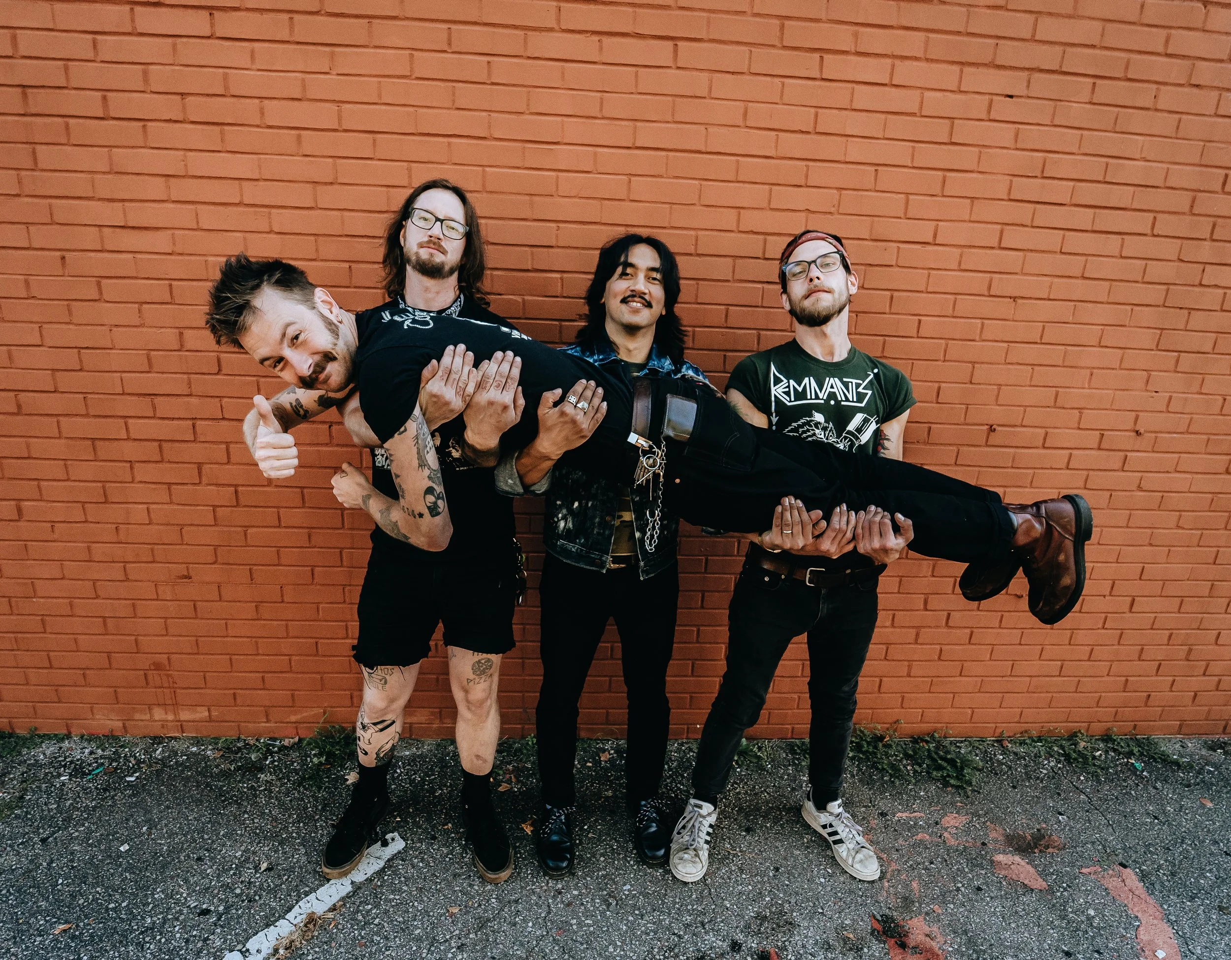 Four young men with tattoos, glasses, and facial hair standing against a red brick wall. One is carrying another horizontally, and they are all smiling or making gestures.