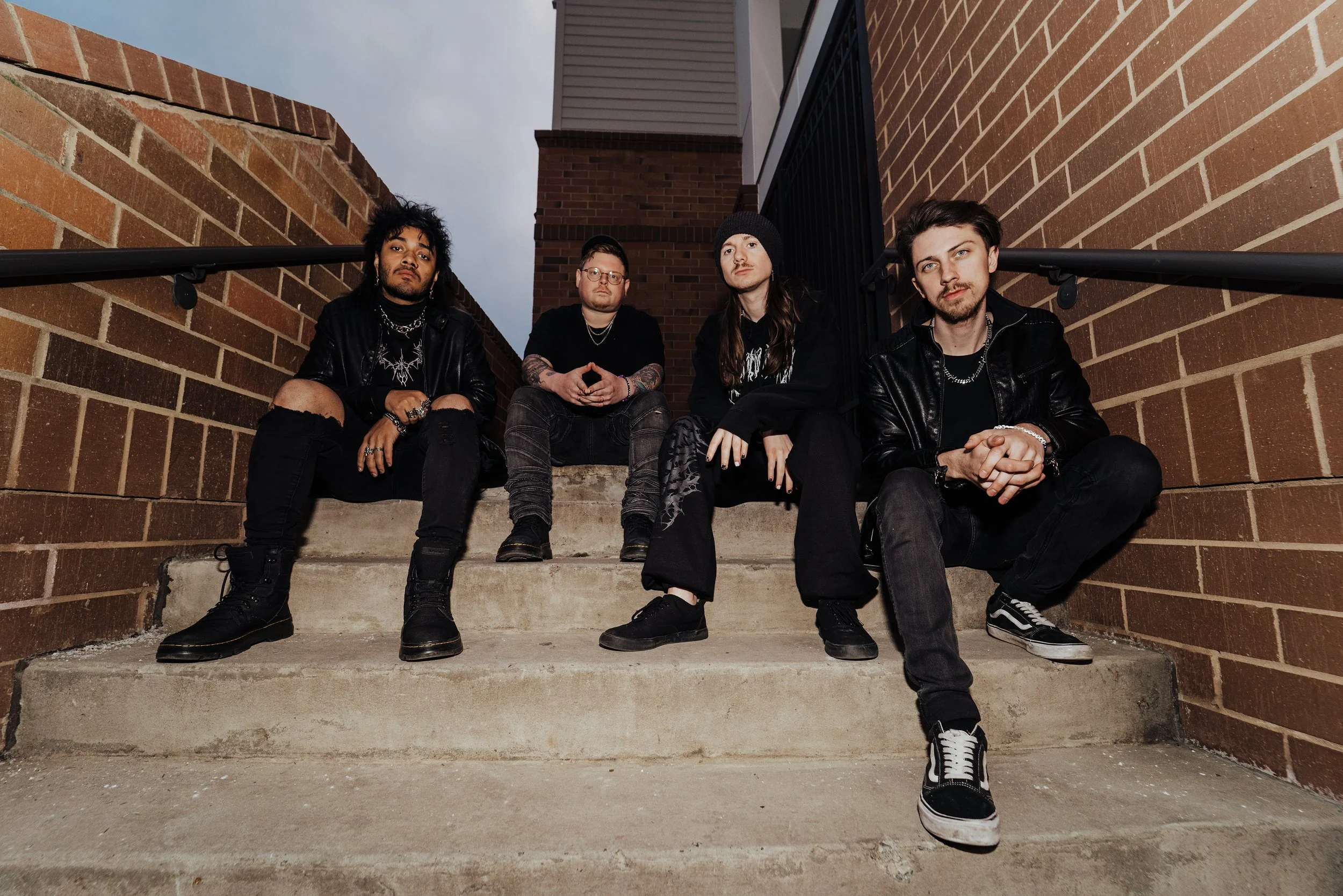 Four young men sitting on concrete steps between brick walls, dressed in black clothing, with a cloudy sky above.