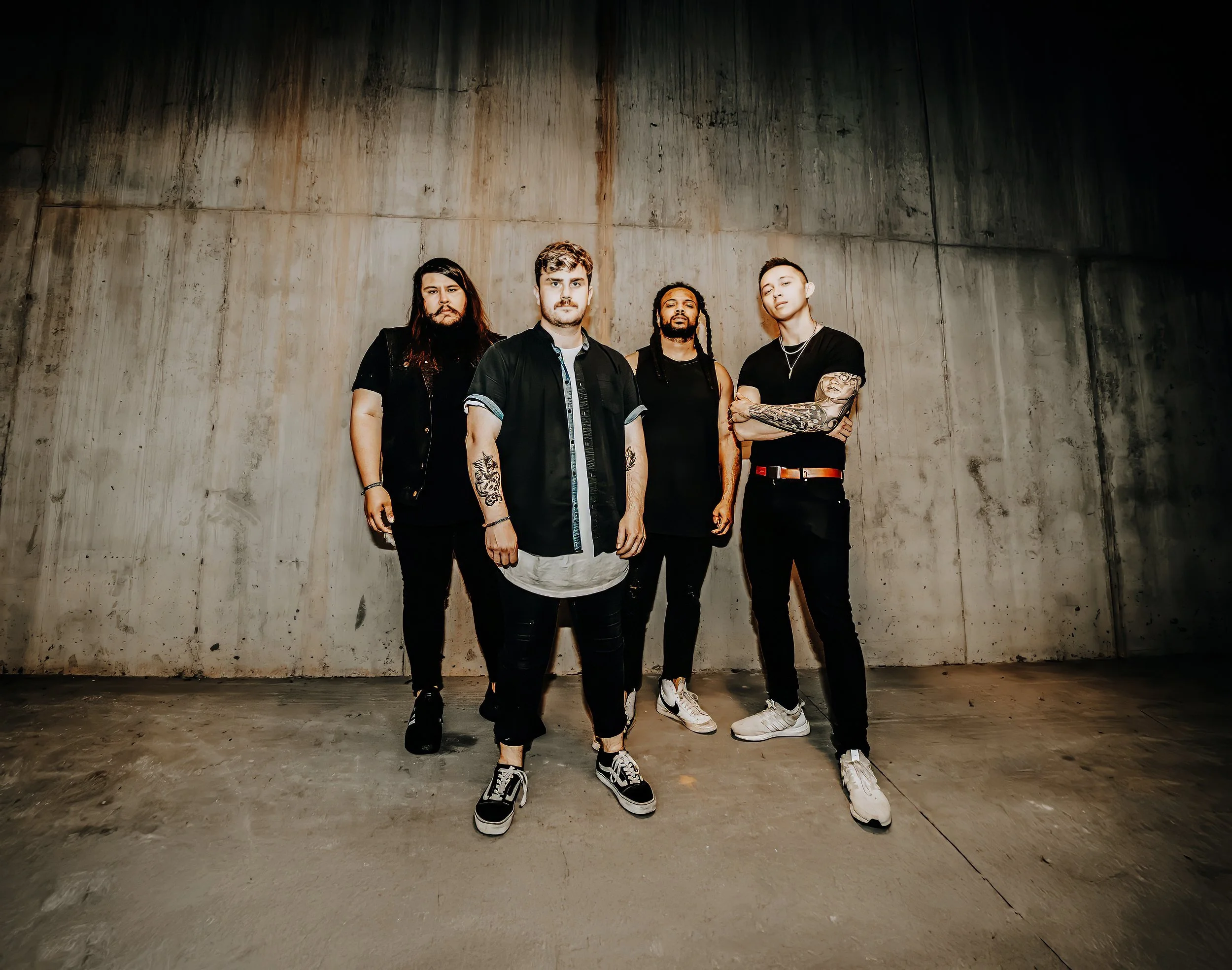Four young men standing in front of a concrete wall indoors, dressed in casual streetwear with tattoos and various hairstyles, posing confidently for the photo.
