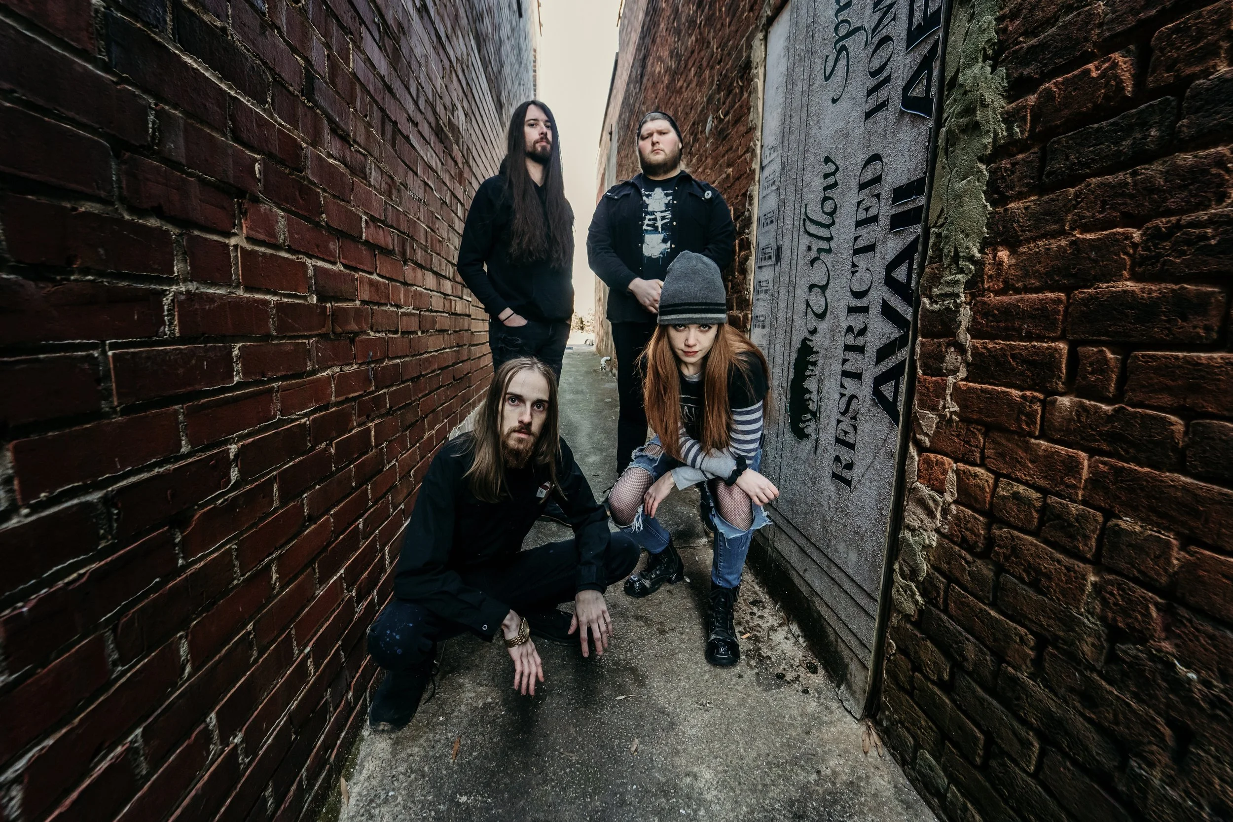 Four young people with dark hair and casual clothing in a narrow alley between brick walls, with one crouching and the others standing, one wearing a beanie and others with long hair, some with serious expressions.