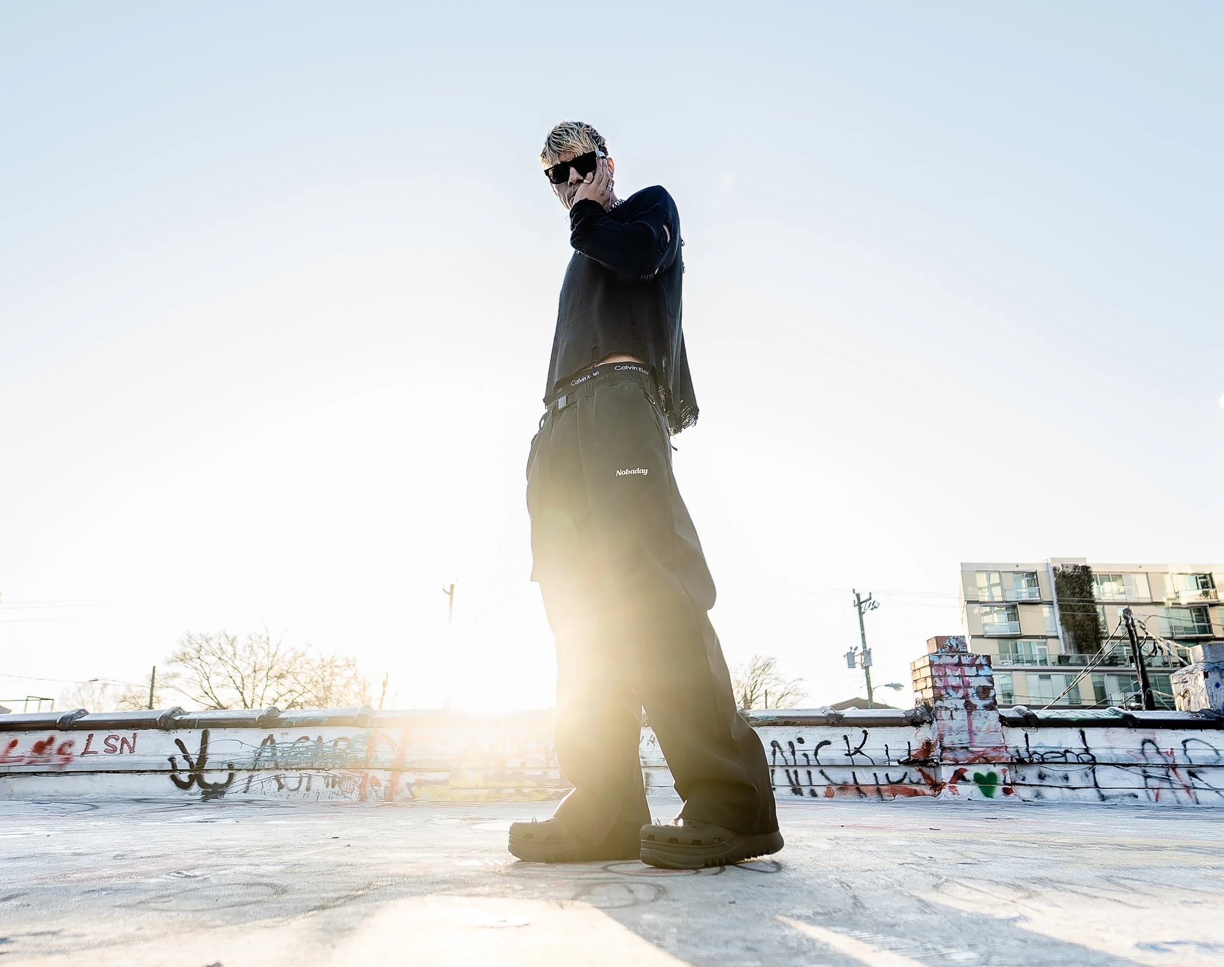 A person standing outdoors on a bright, sunny day, wearing sunglasses and a black jacket, with graffiti-covered wall in the foreground and buildings in the background.