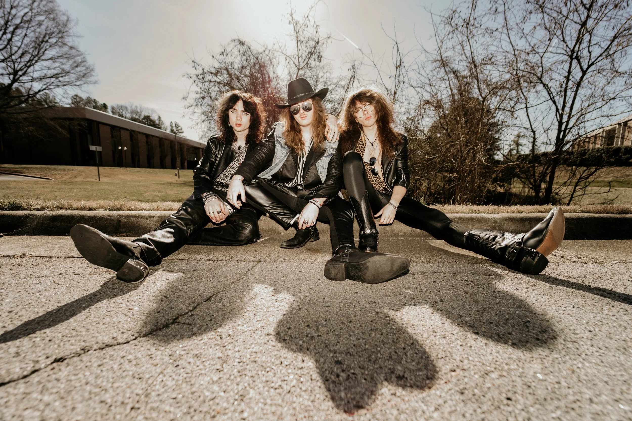 Three young women sitting on a street curb during daytime, dressed in rocker style clothing with leather jackets, sunglasses, and tousled hair, against a background of leafless trees and a clear sky.