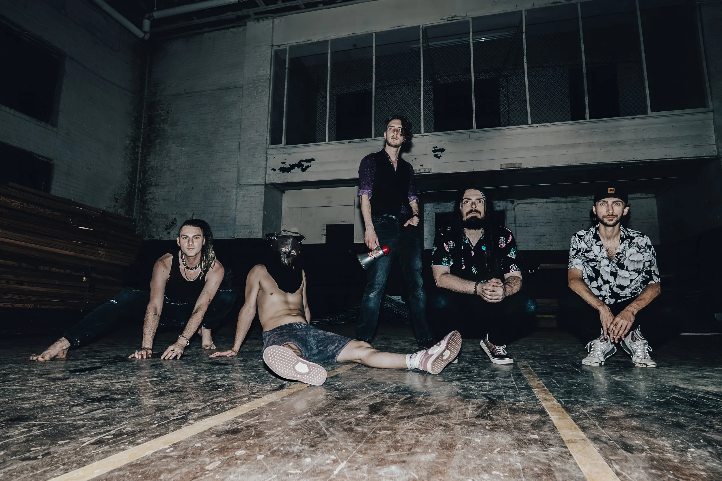 Group of five young men, four sitting and one standing, in an industrial-style indoor space with a distressed wall and large windows in the background. Two of them are shirtless, one is wearing a patterned shirt and a cap, and another is holding a me