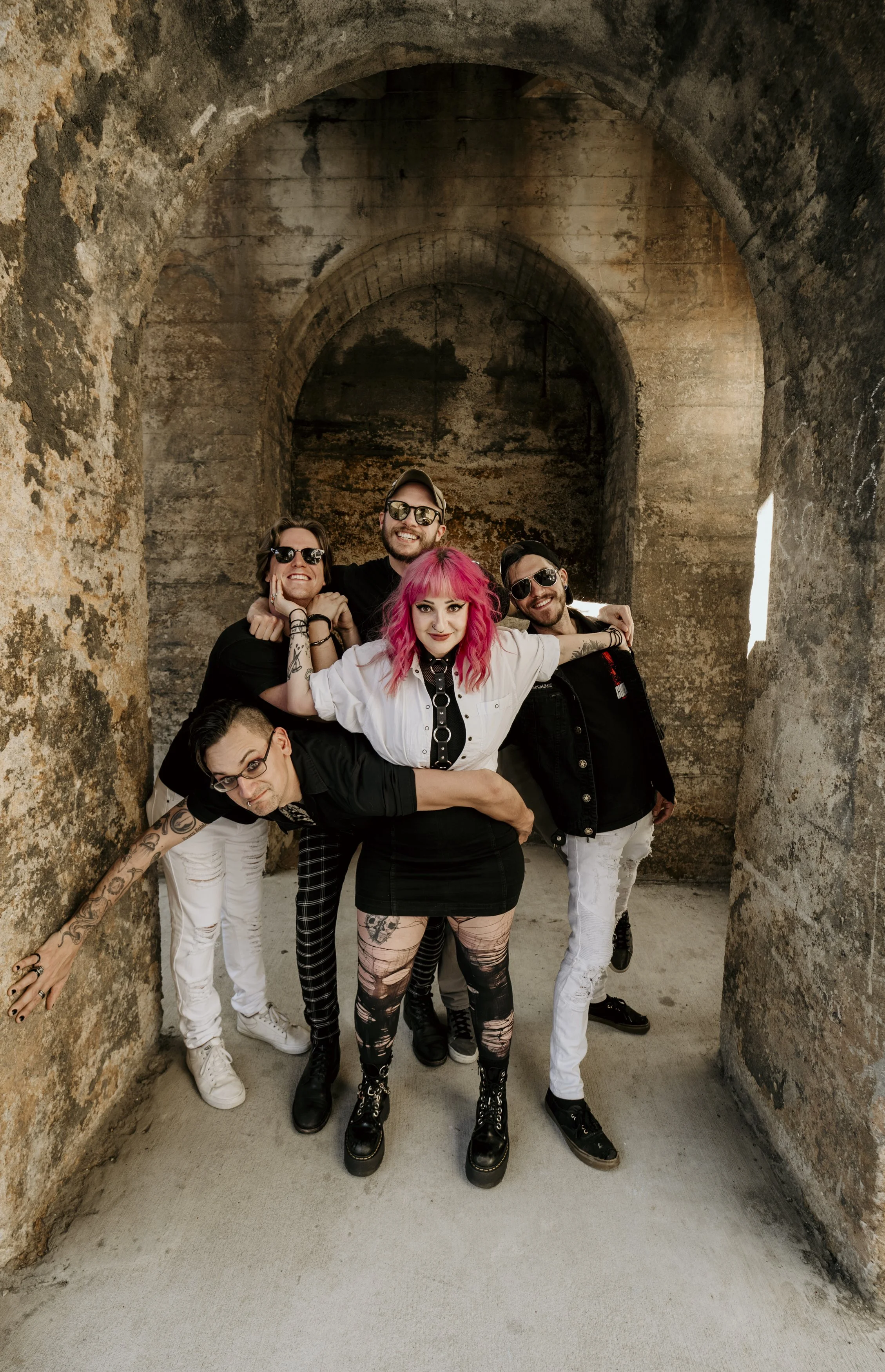 A group of six young adults posing inside an old, arched stone structure with textured walls. They are dressed in alternative fashion style, with some wearing sunglasses, and are smiling or making playful expressions.