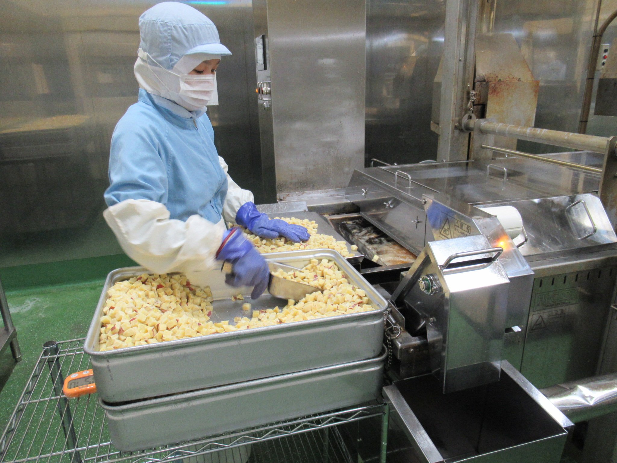 白い帽子とマスクをつけた作業者が工場のラインで食品をカットしている。ステンレスの作業台と機械が見える。