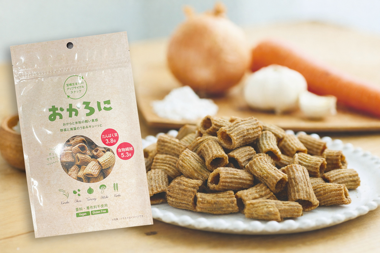 ベージュの袋に緑色の文字と画像があり、里芋の形をした野菜スナックのお菓子のパッケージ。背景に里芋、にんじん、にんにくが置いてある木の板と白い皿に盛られた野菜のスナックの写真。