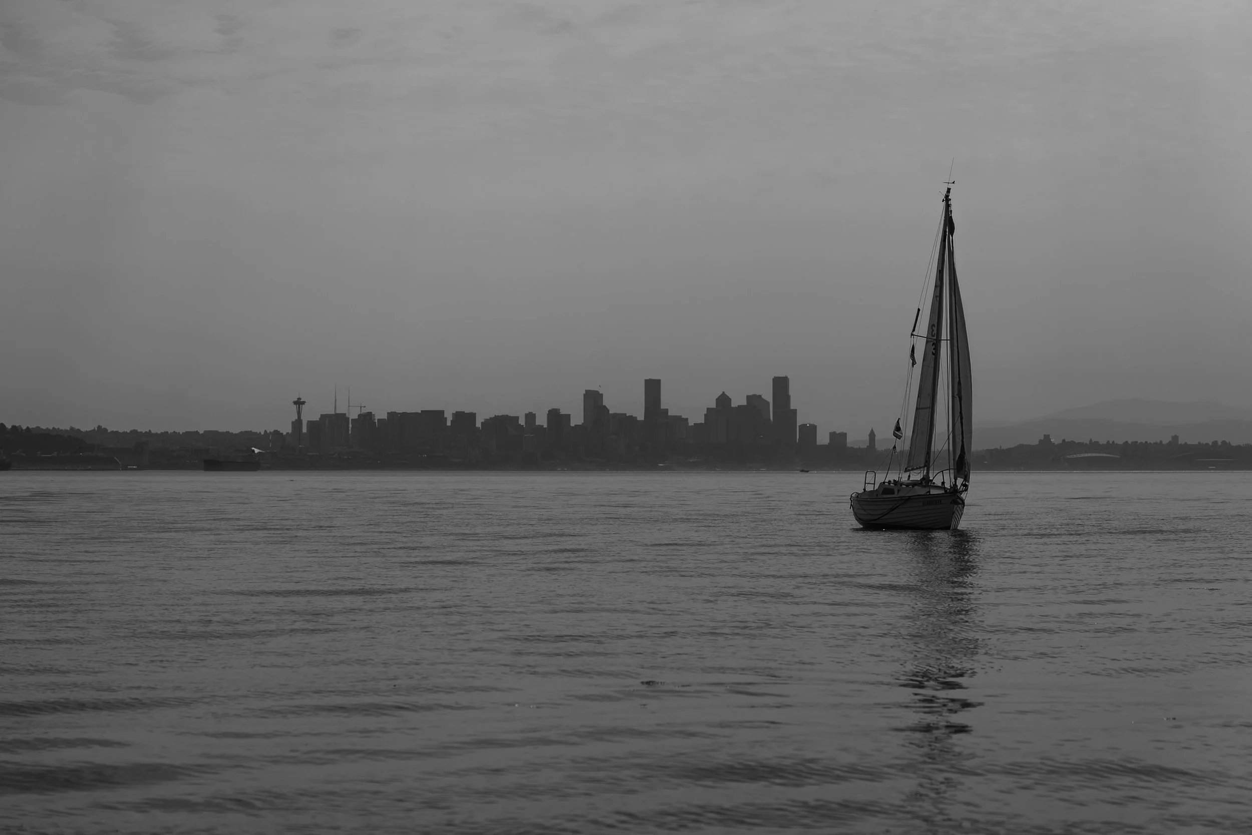 A sailboat on calm water with the Seattle skyline and the Space Needle in the background, under a cloudy sky.