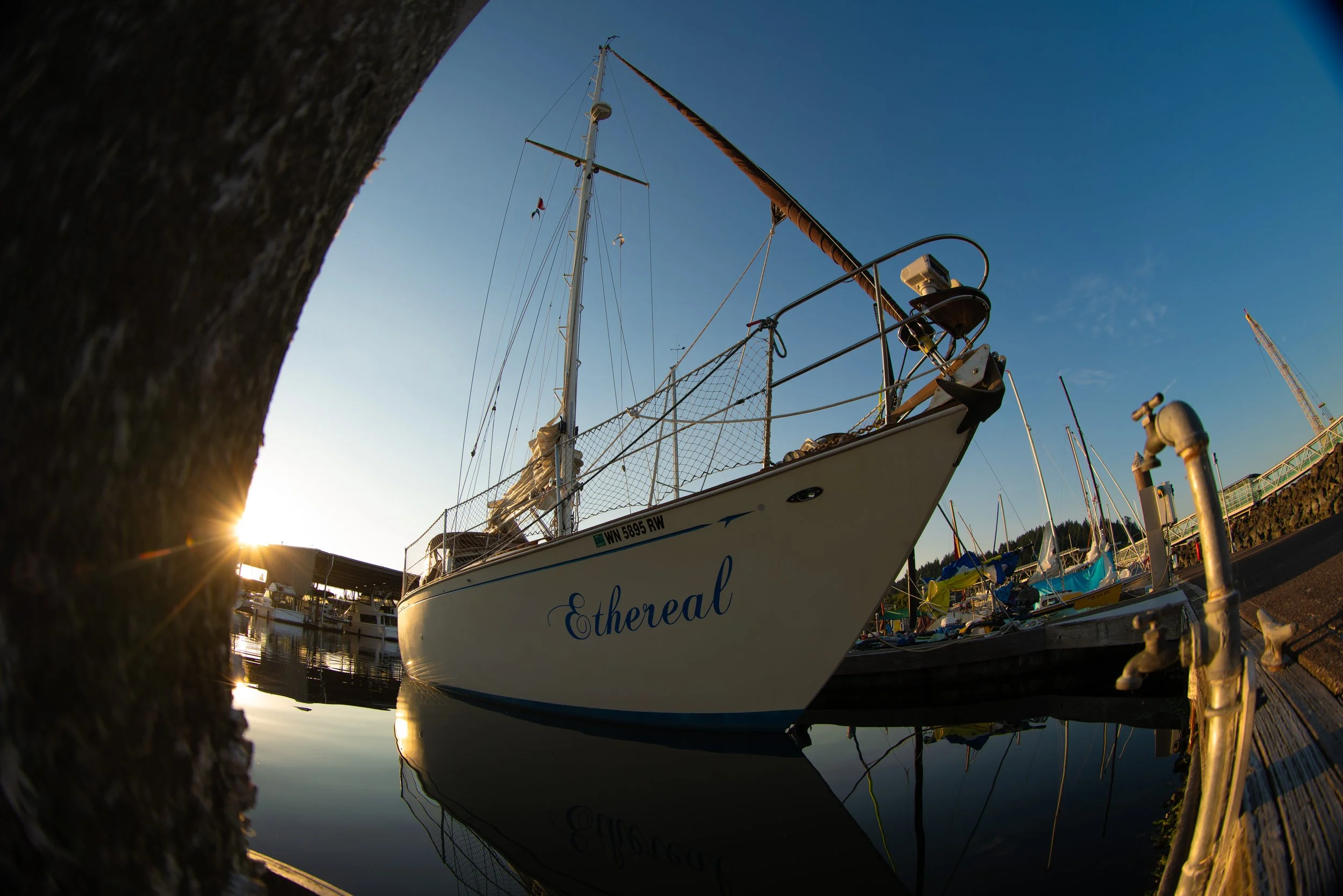 View of a sailboat named Ethereal docked at a marina during sunset. The photo is taken from a low angle, with the boat's reflection visible in the calm water. The sky is clear and blue, and a crane is visible in the background.