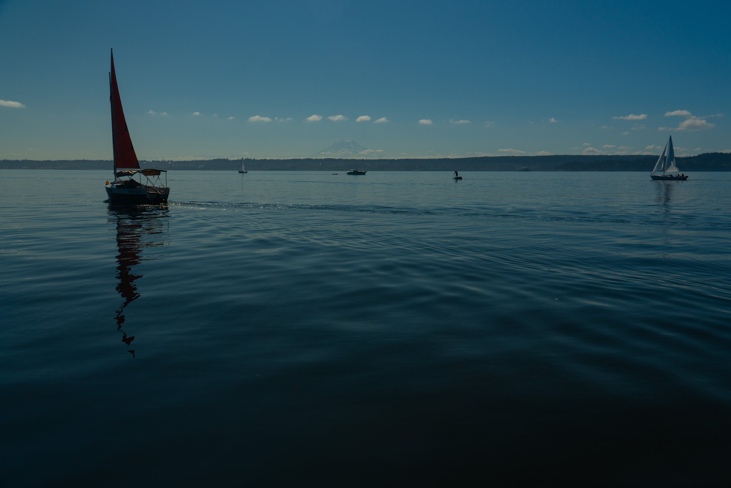 Calm water with several sailboats and a motorboat, with mountains and a partly cloudy sky in the background.