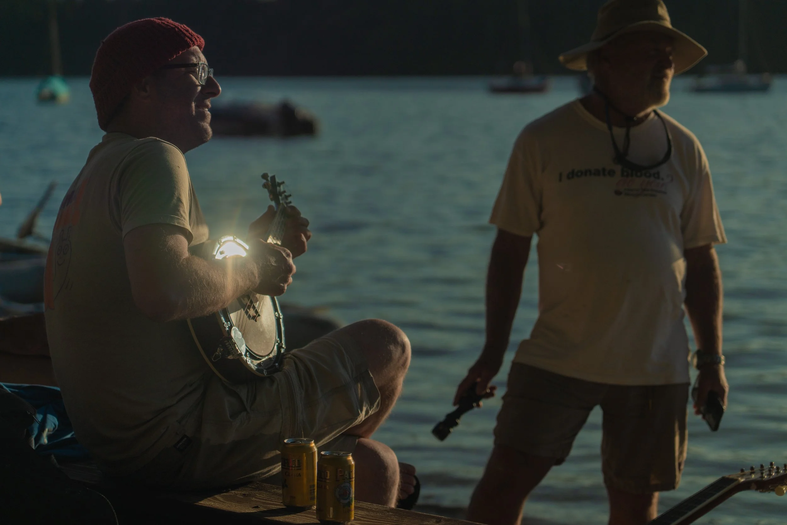 Two men sitting by a body of water, one playing a guitar and the other standing nearby, with boats in the background during sunset.