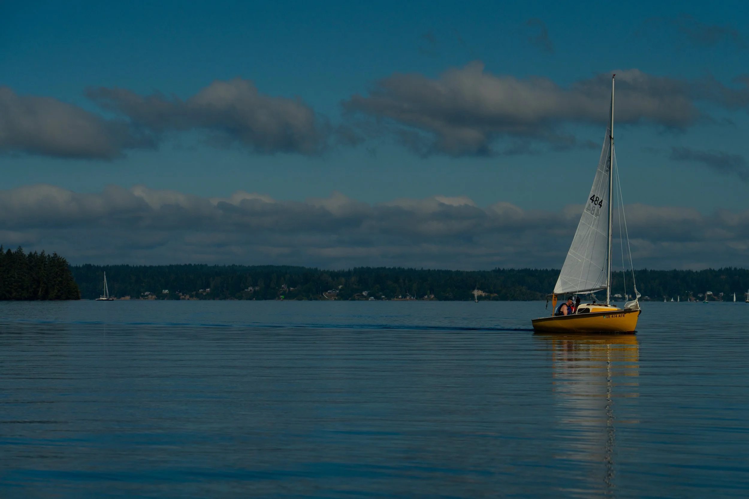A yellow sailboat with two people on board sailing on a calm body of water, with a forested shoreline and a few other sailboats in the background under a mostly cloudy sky.