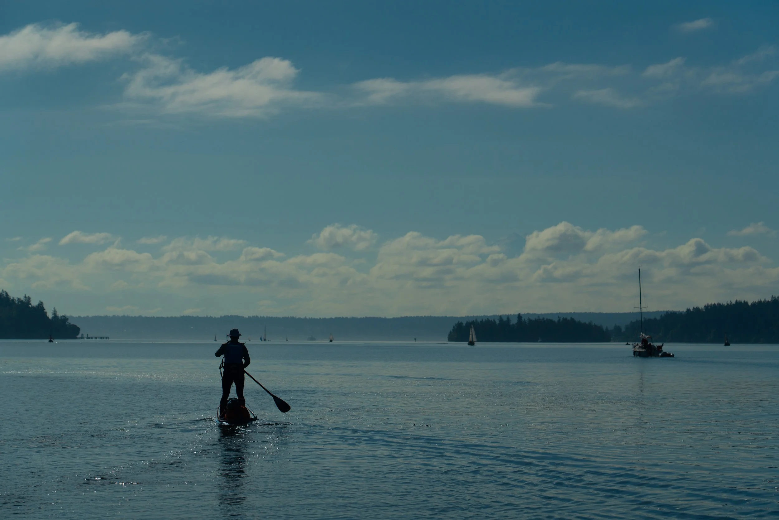 A person paddleboarding on a lake with sailboats and forested land in the background, under a partly cloudy sky.
