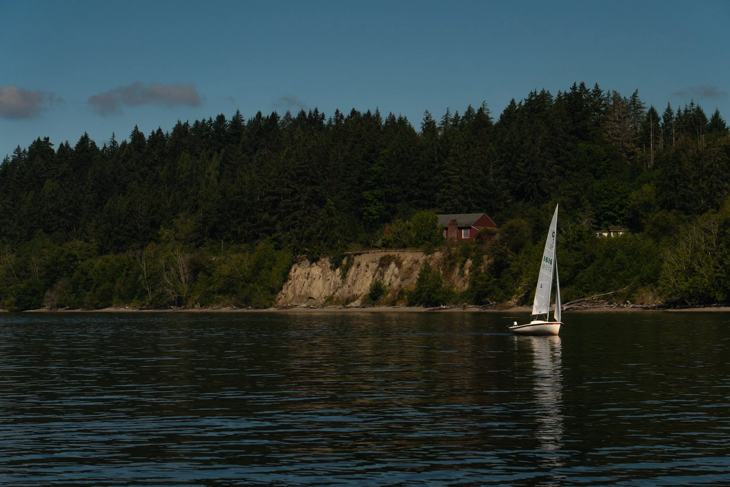 A sailboat with a white sail on a body of water, with a forested hillside and houses in the background, under a clear blue sky with some clouds.