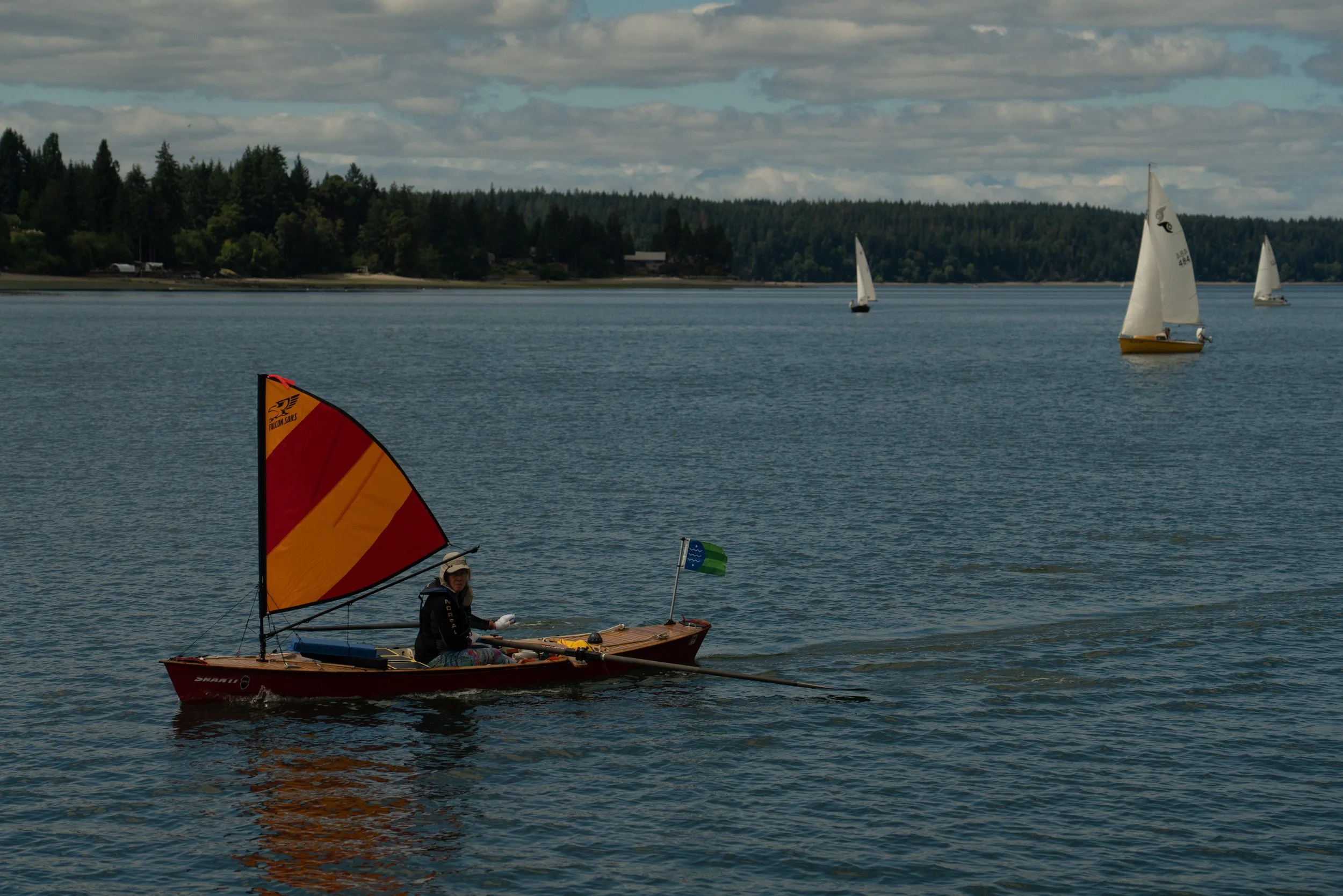 A person sailing a small boat with a bright orange and yellow sail on a lake or river, with a backdrop of tree-lined shoreline and other sailboats in the distance under a partly cloudy sky.