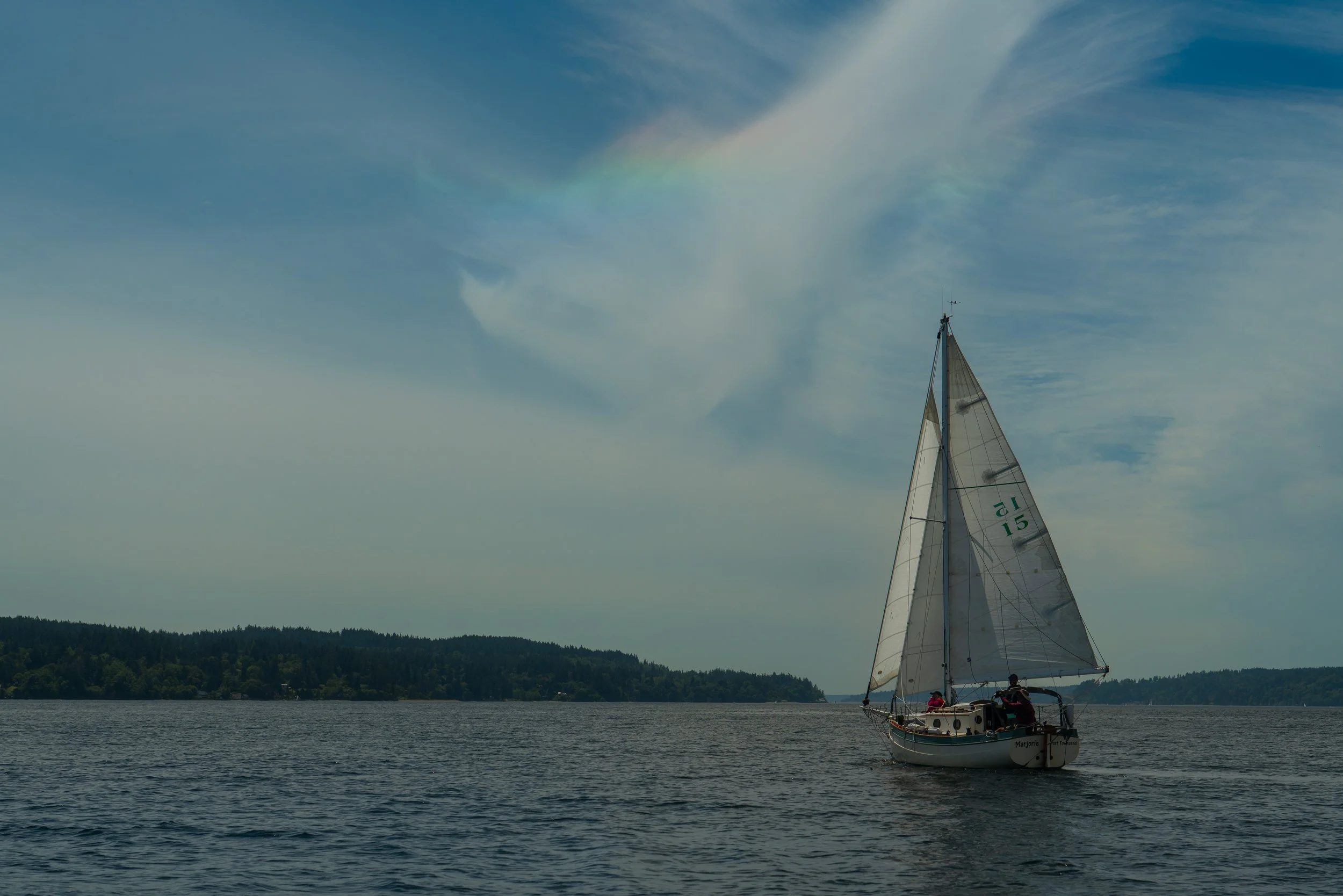 A sailboat with white sails on a body of water under a partly cloudy sky. There is a faint rainbow visible in the clouds. The shoreline with trees is visible in the background.