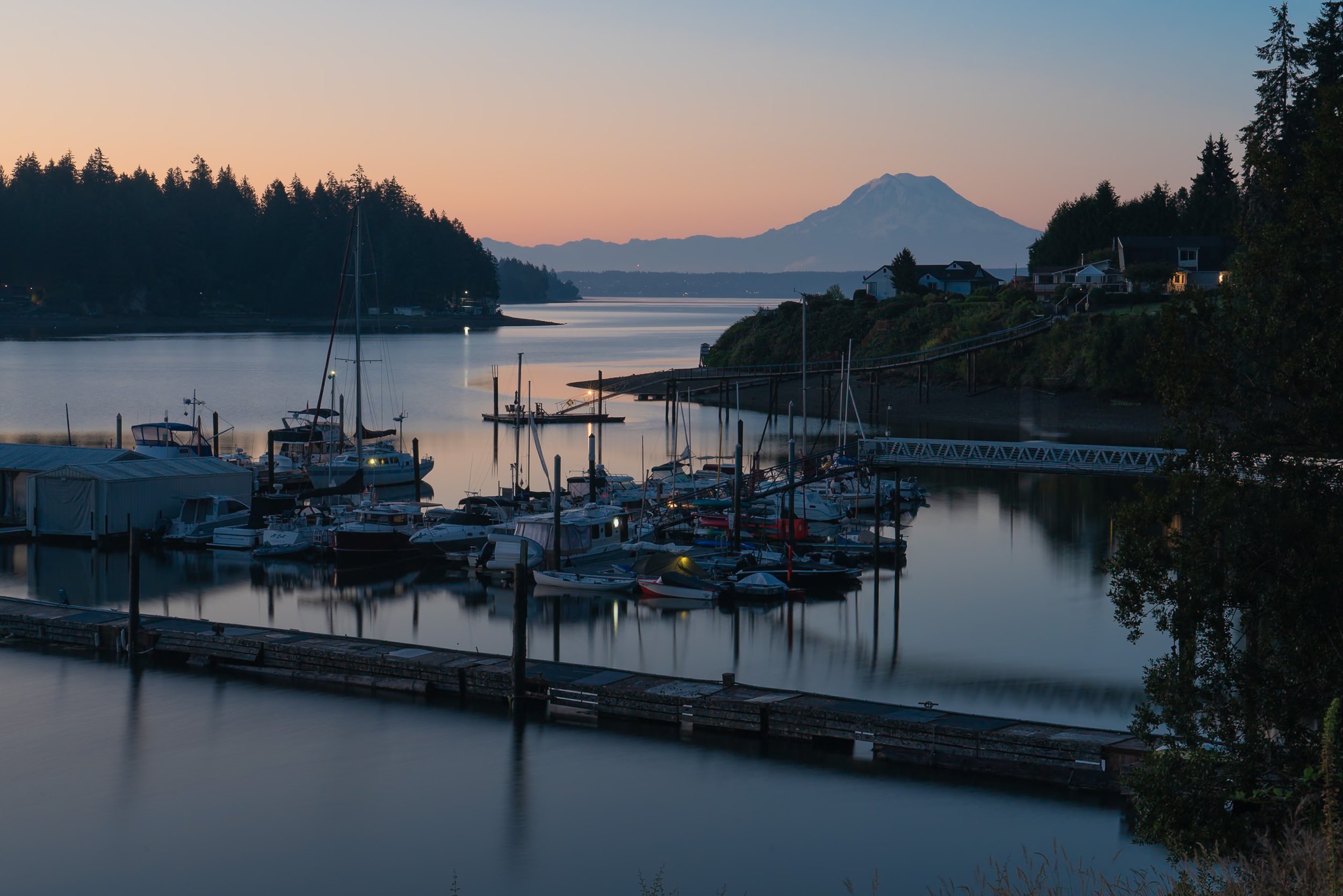 A marina at dusk with boats docked, calm water reflecting the sky, and a mountain in the background under a pastel-colored sky.