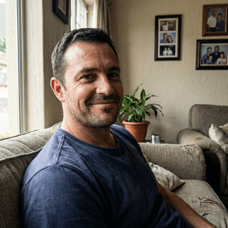 A young man comforting an elderly man on a sofa in a cozy living room, holding hands lovingly, with family photos on the wall behind them.